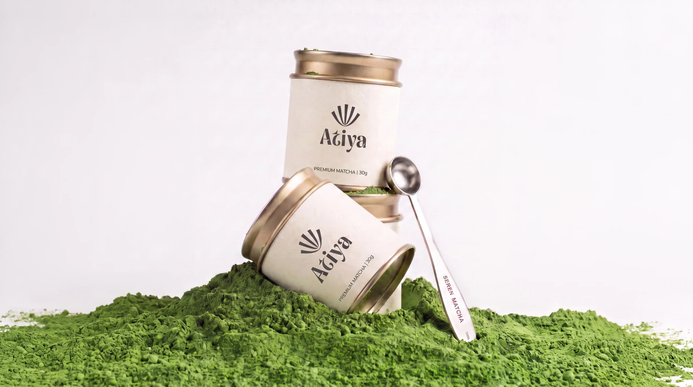 Two Atiya tins, one upright and one leaning, standing on a mound of loose matcha powder with a long measuring spoon