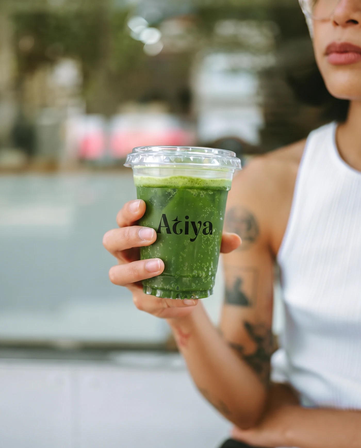 A woman on a street holding an Atiya cup of iced matcha, tattooed arm, white vest