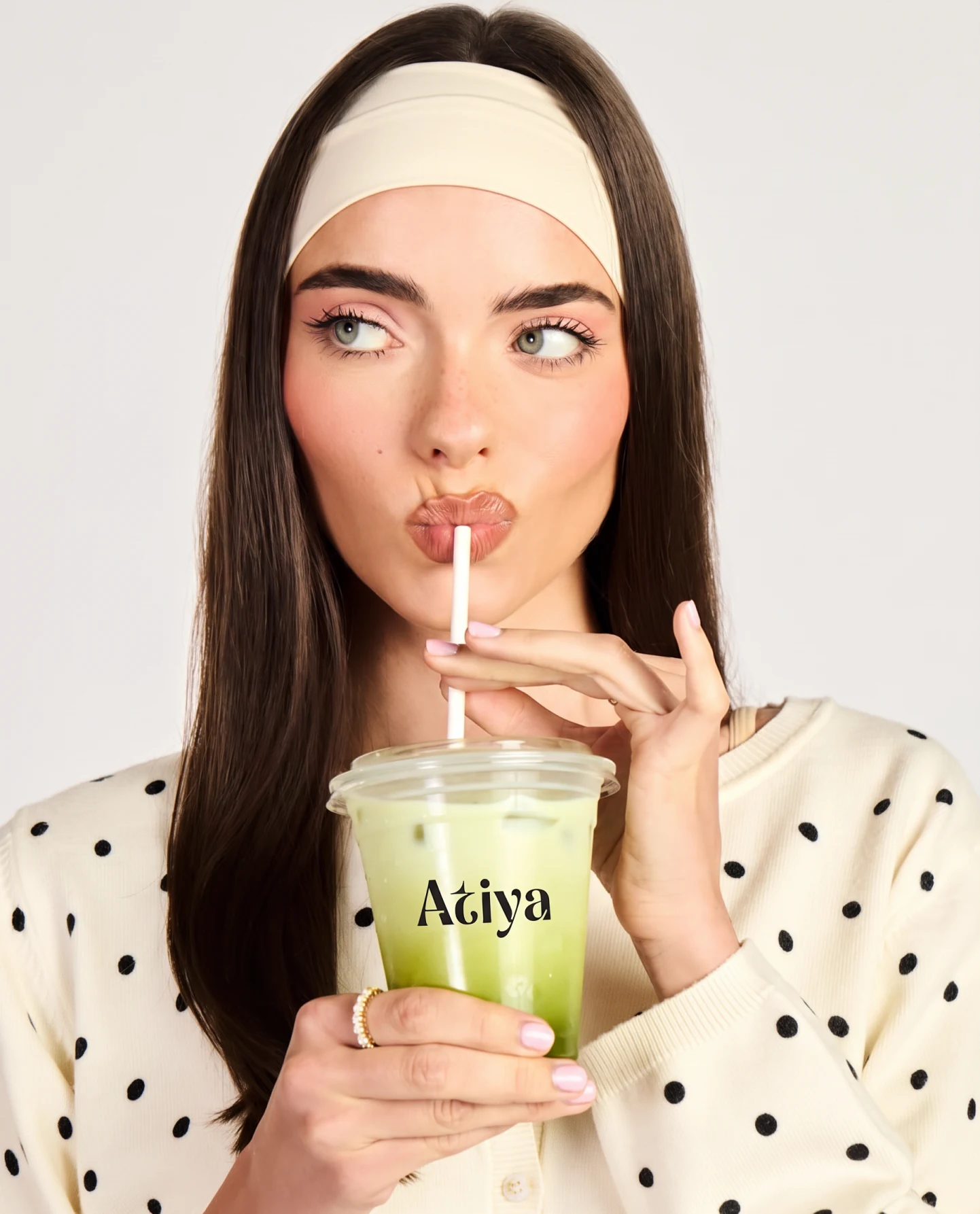 A woman in a polka-dot shirt with a headband, drinking iced matcha through a straw from an Atiya cup