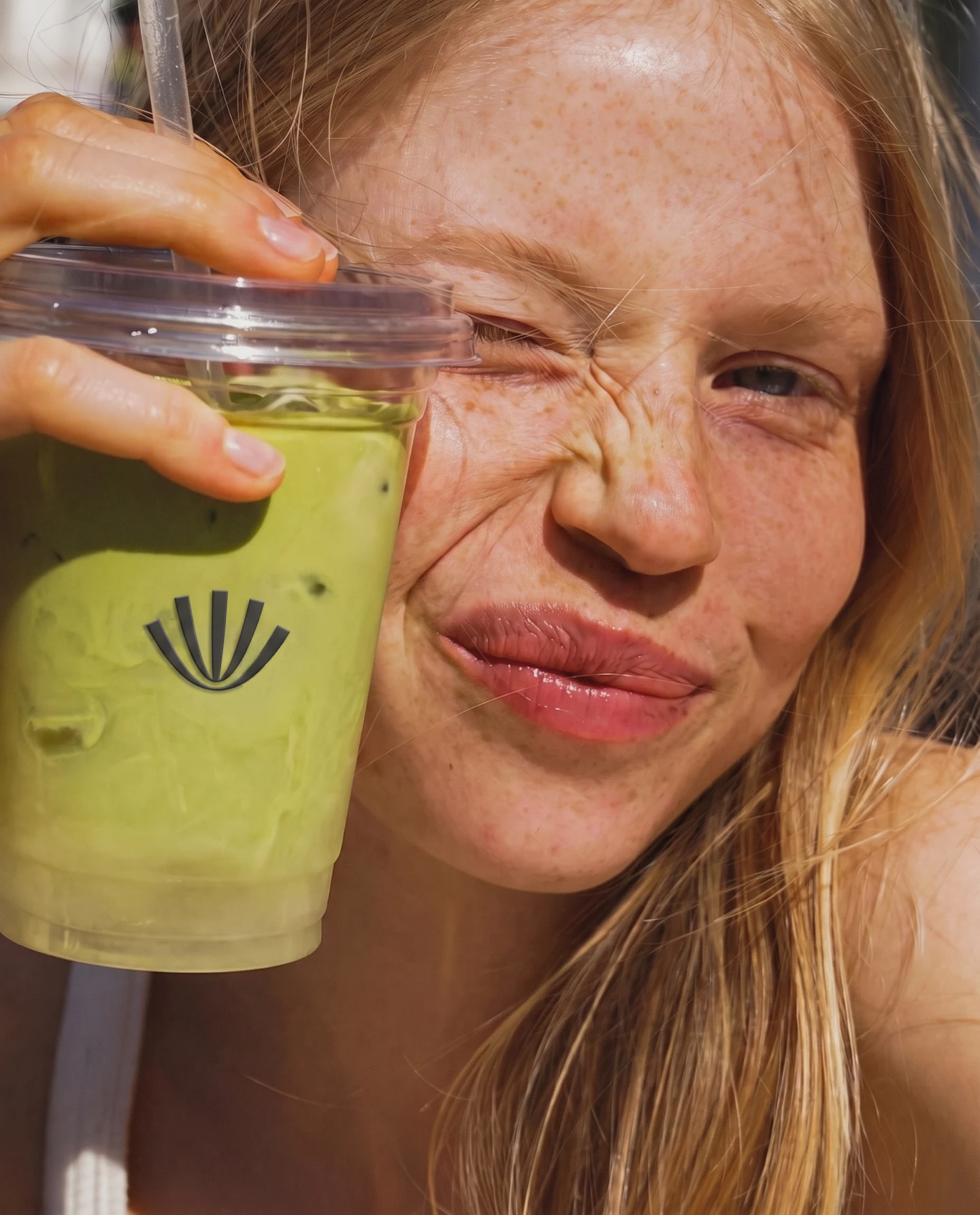 A woman holding a cup of matcha up beside her face, the Atiya whisk symbol printed on the cup