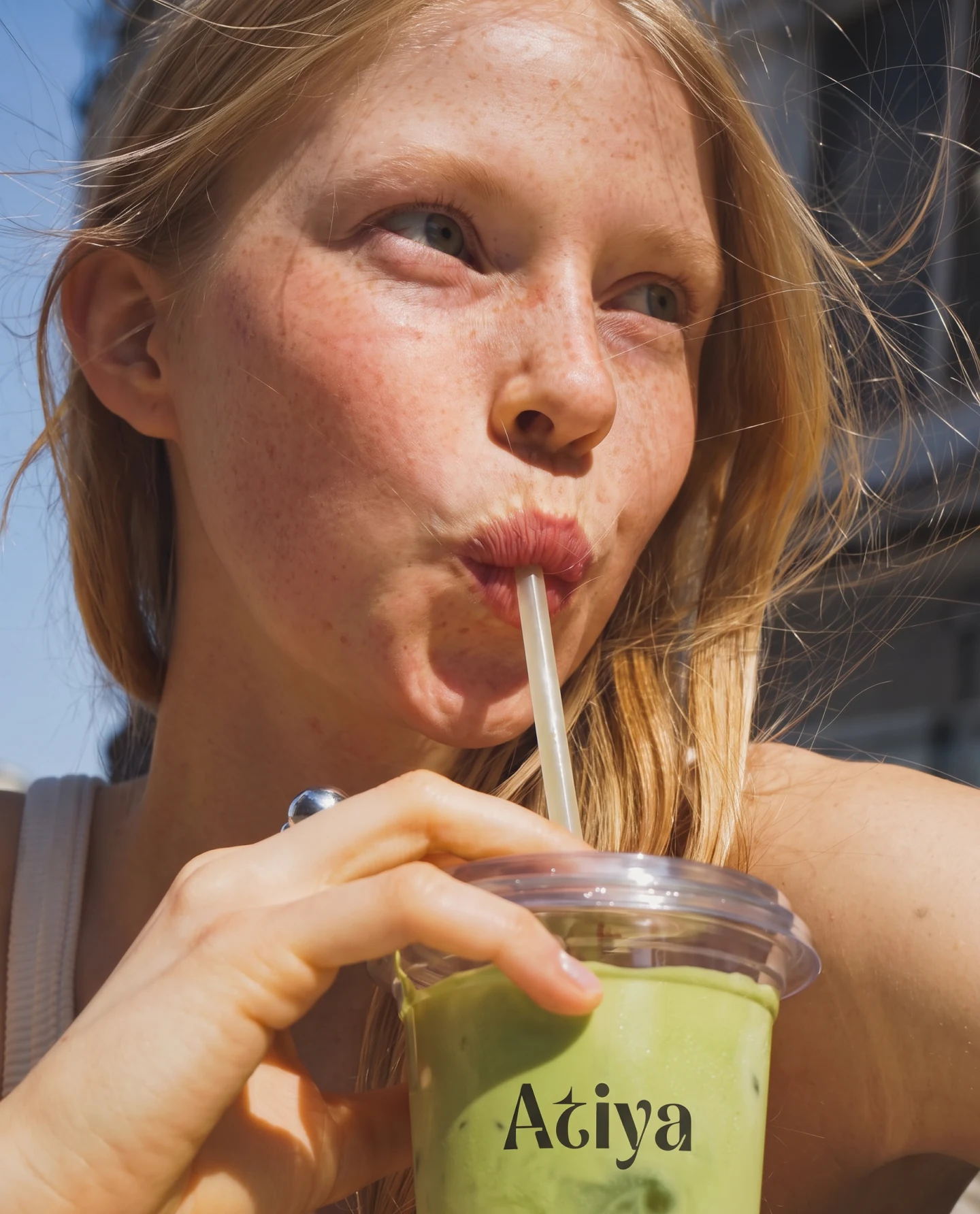 A woman drinking iced matcha through a straw from an Atiya branded cup, shot close in daylight