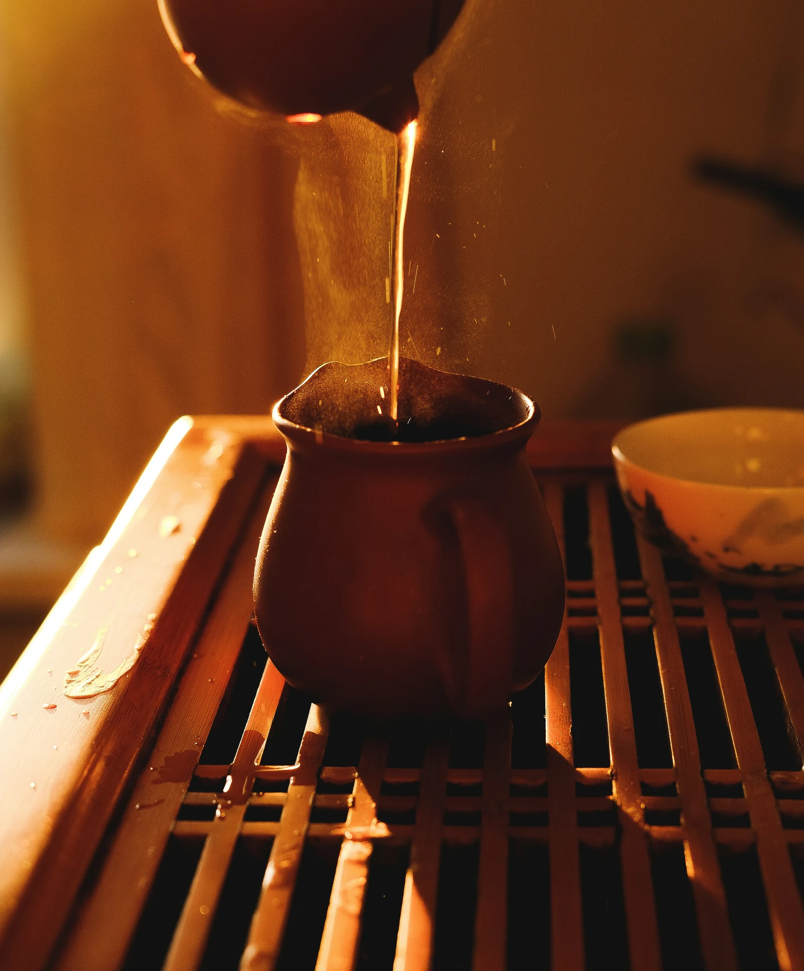 Tea poured from a clay pot into a small cup on a wooden tea tray, in warm lamplight