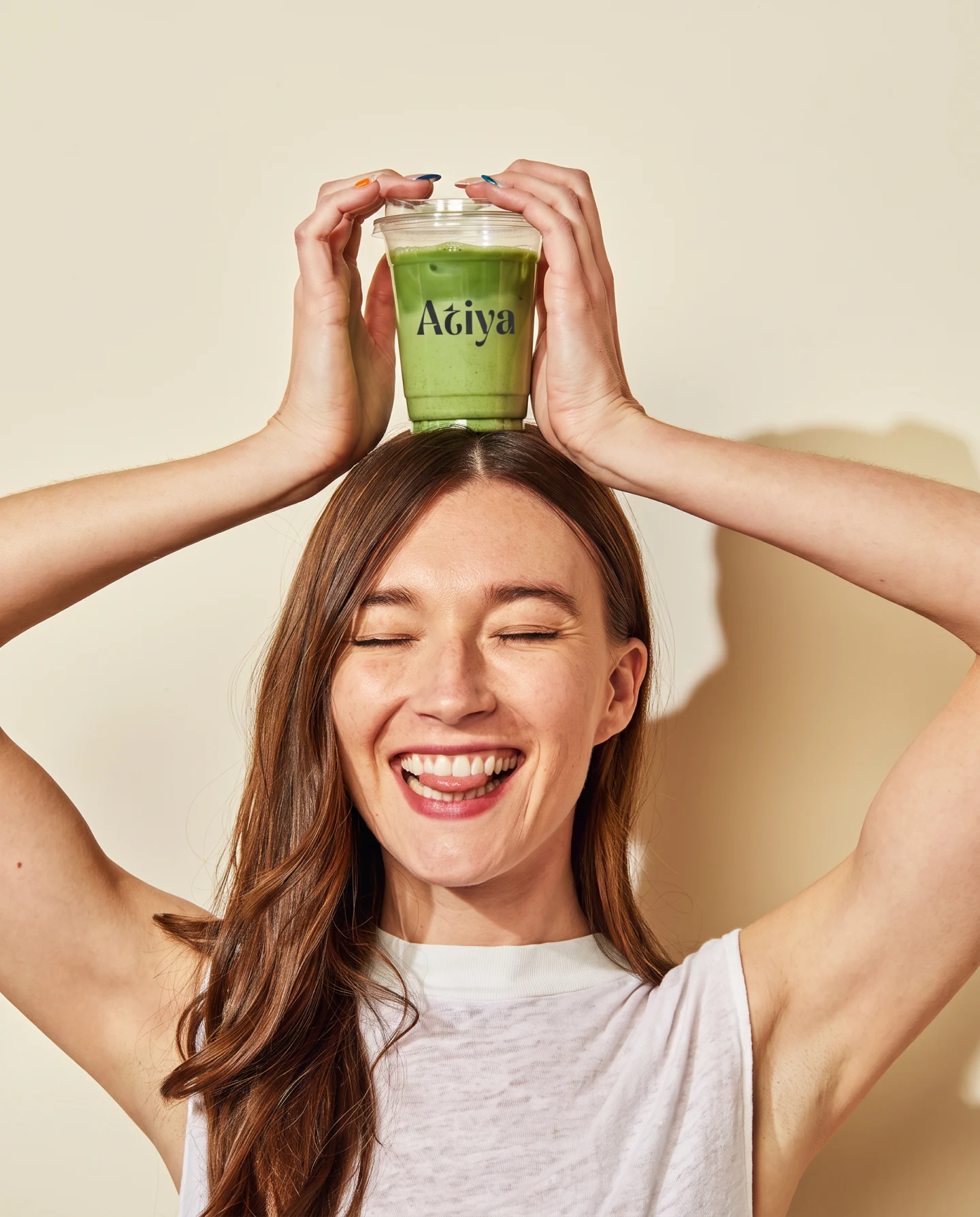 A woman holding an Atiya cup above her head with both hands, laughing, on a cream ground