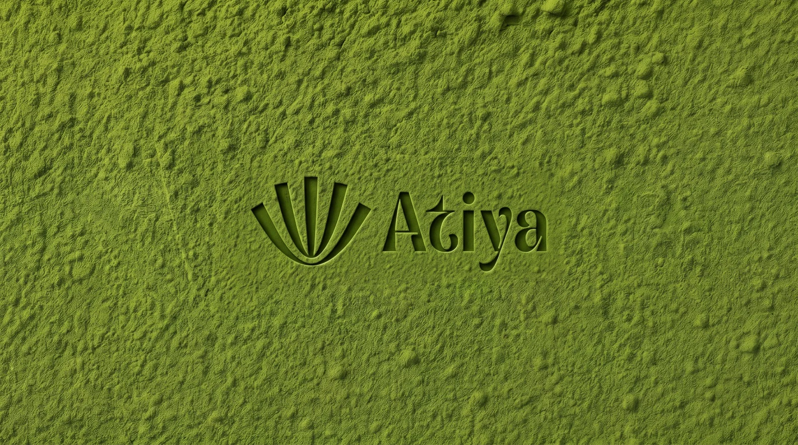 The Atiya symbol and wordmark pressed into a bed of sifted matcha powder, the debossed edges catching a low raking light