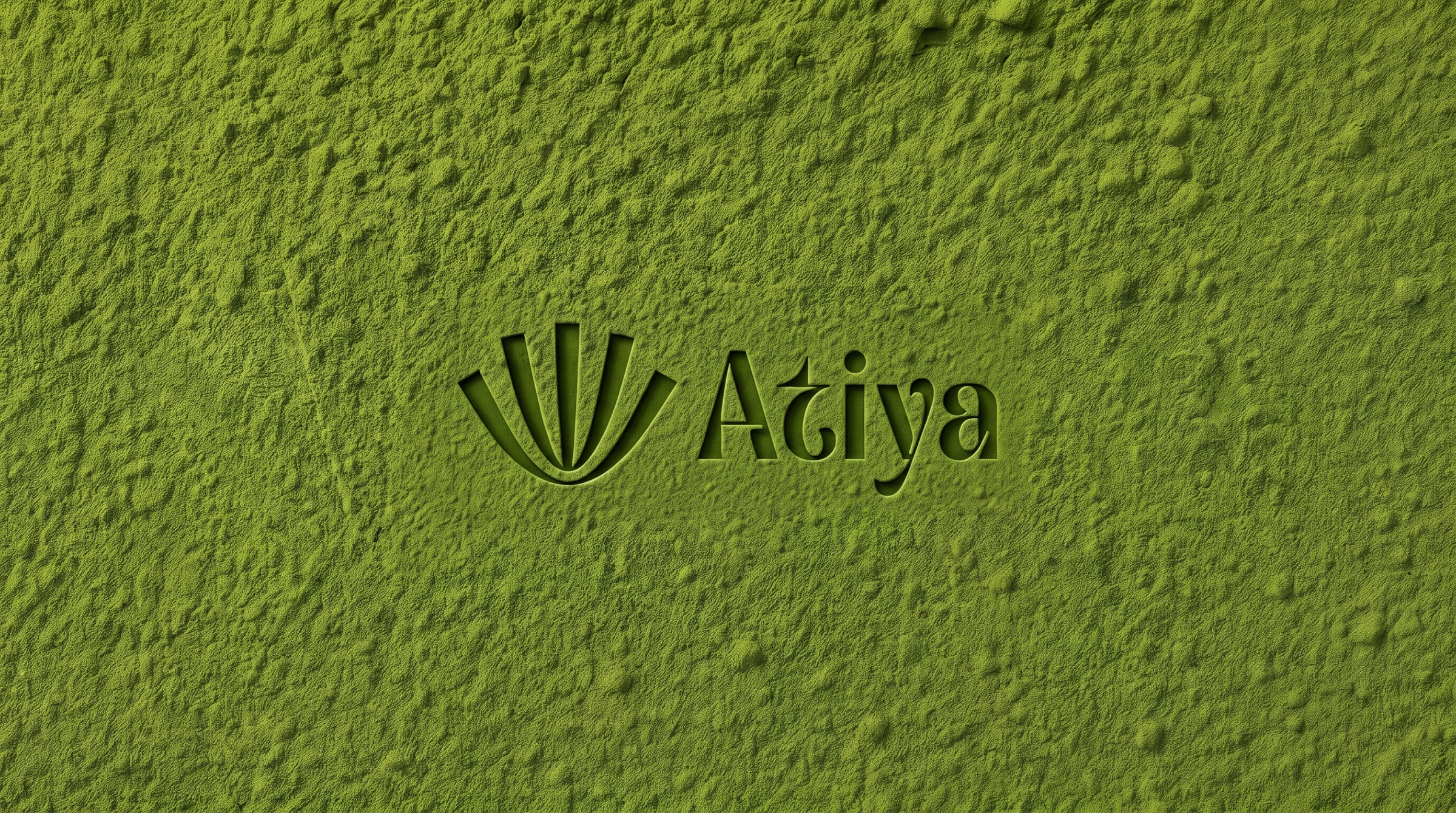 The Atiya symbol and wordmark pressed into a bed of sifted matcha powder, the debossed edges catching a low raking light