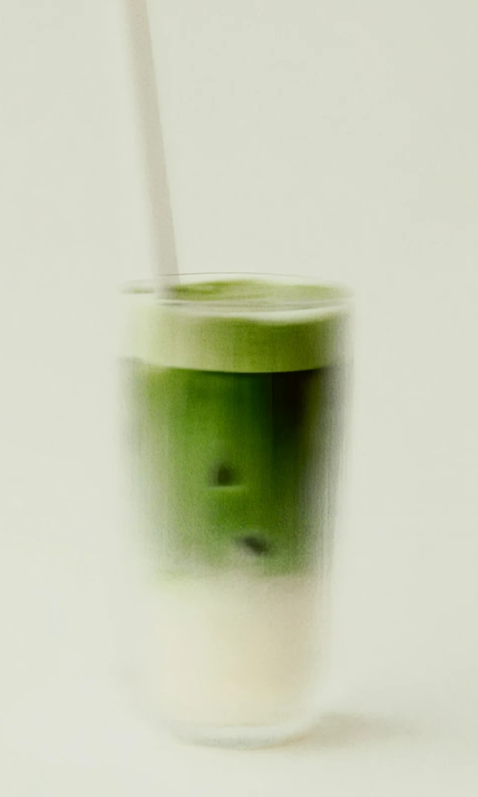 A tall glass of matcha layered over milk with a straw, on a plain pale ground