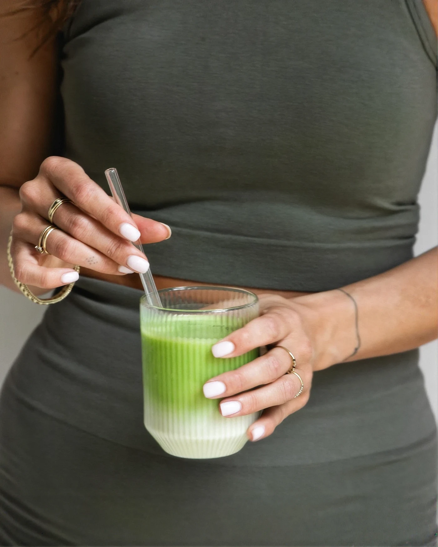 A woman in a grey dress holding a ribbed glass of matcha latte with a glass straw