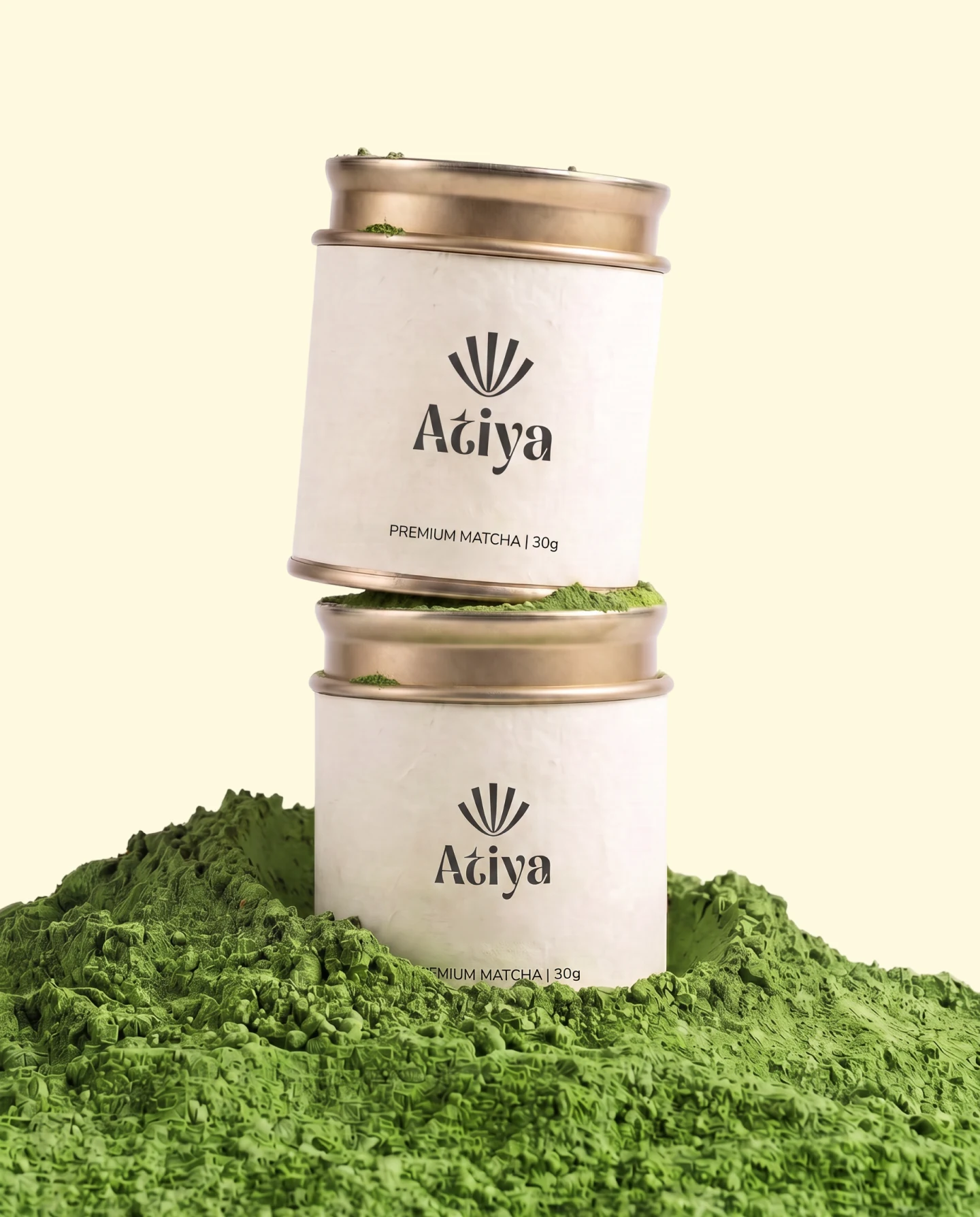 Two Atiya tins stacked on a mound of matcha powder, the gold lids catching the light