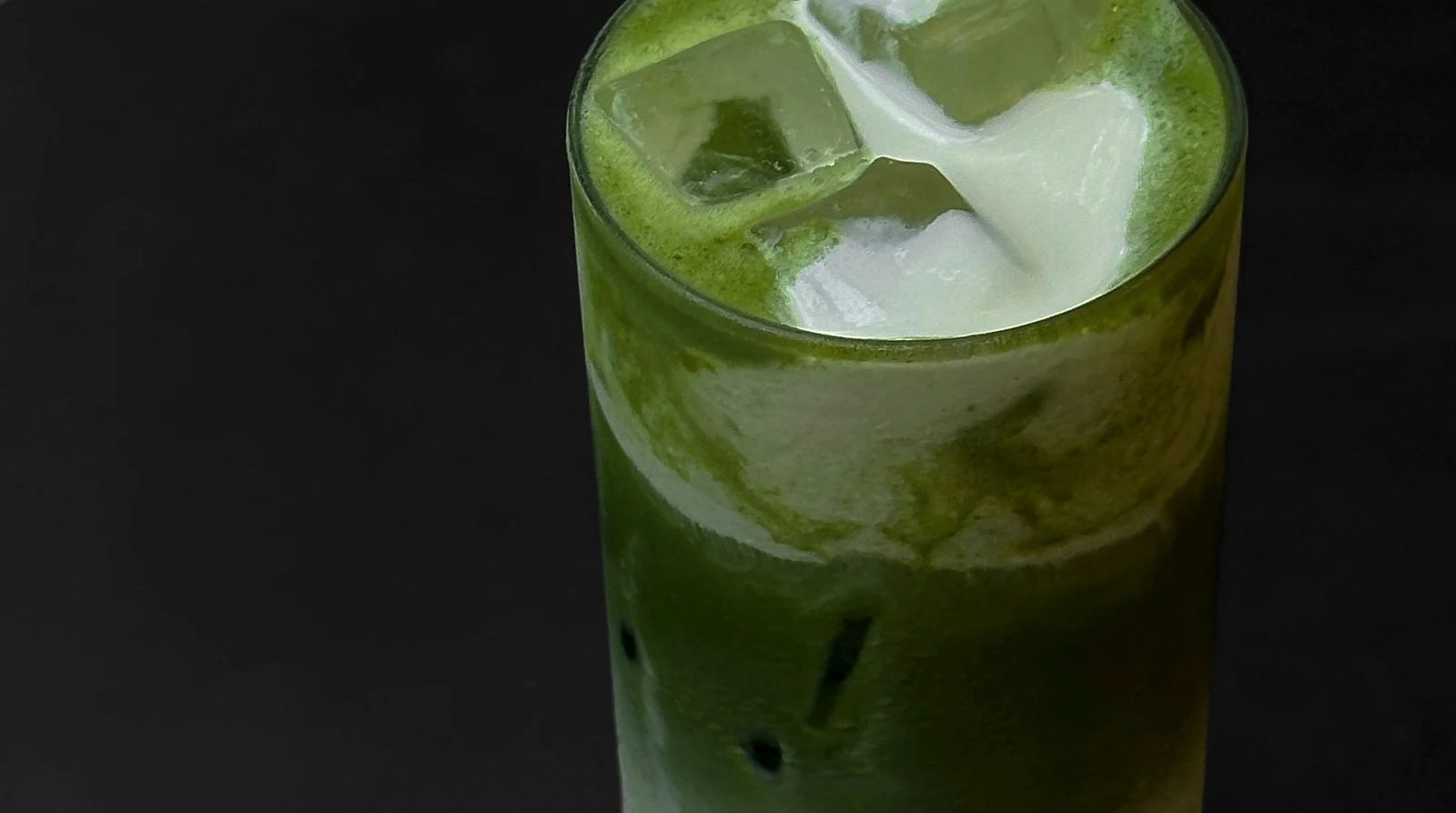 A tall glass of iced matcha layered over milk with a glass straw, on a black table