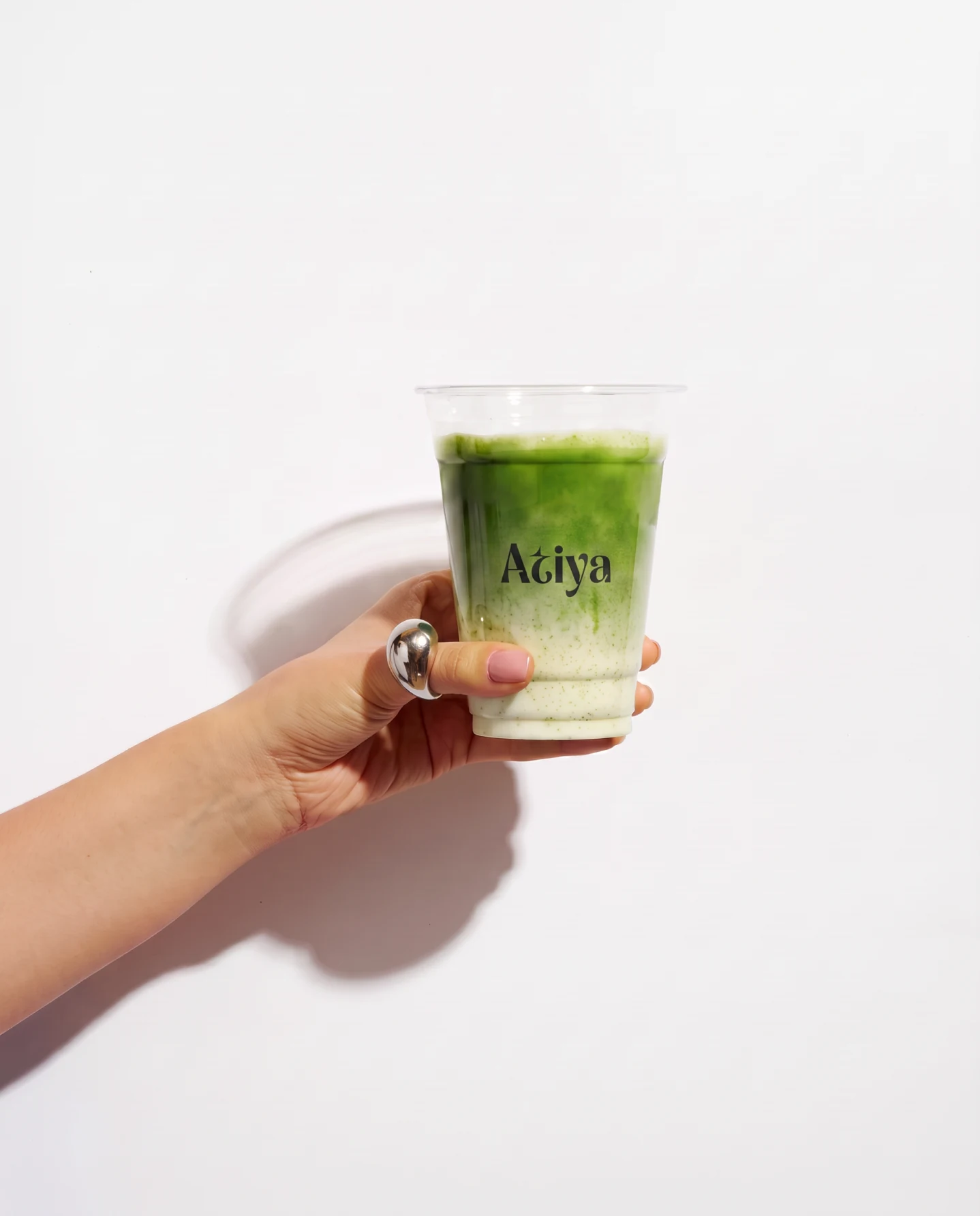 A hand holding a clear cup of iced matcha with the Atiya wordmark on the side, against a plain wall