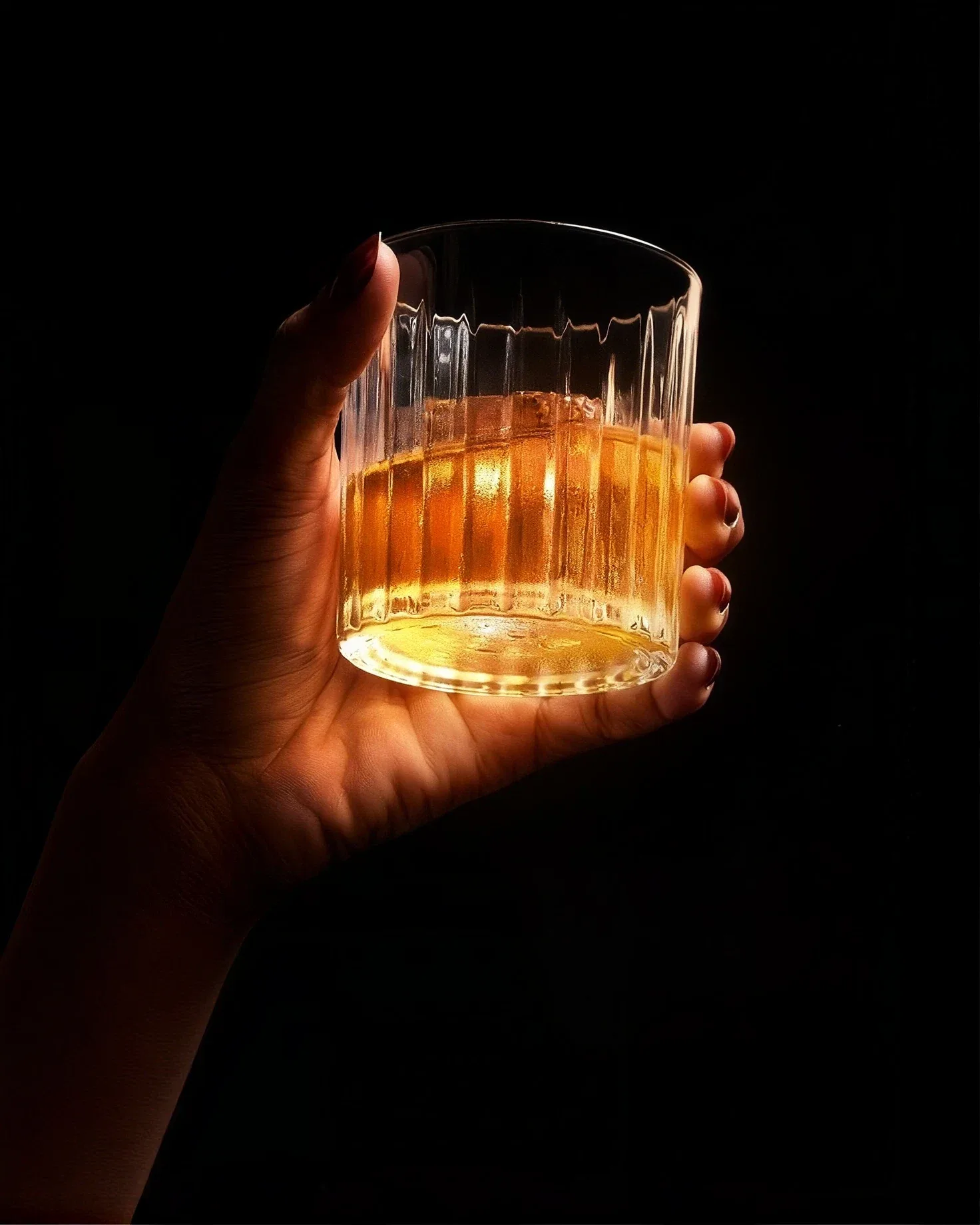 A hand holding a fluted glass of amber spirit, lit against black