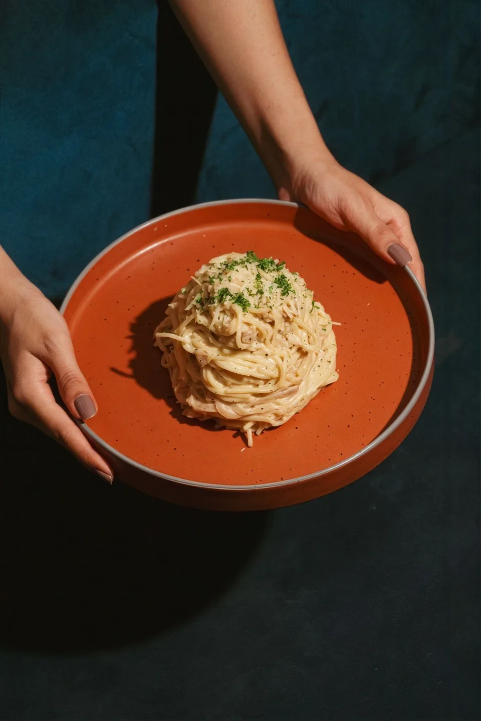 Two hands holding a burnt orange plate of pasta above a deep teal cloth