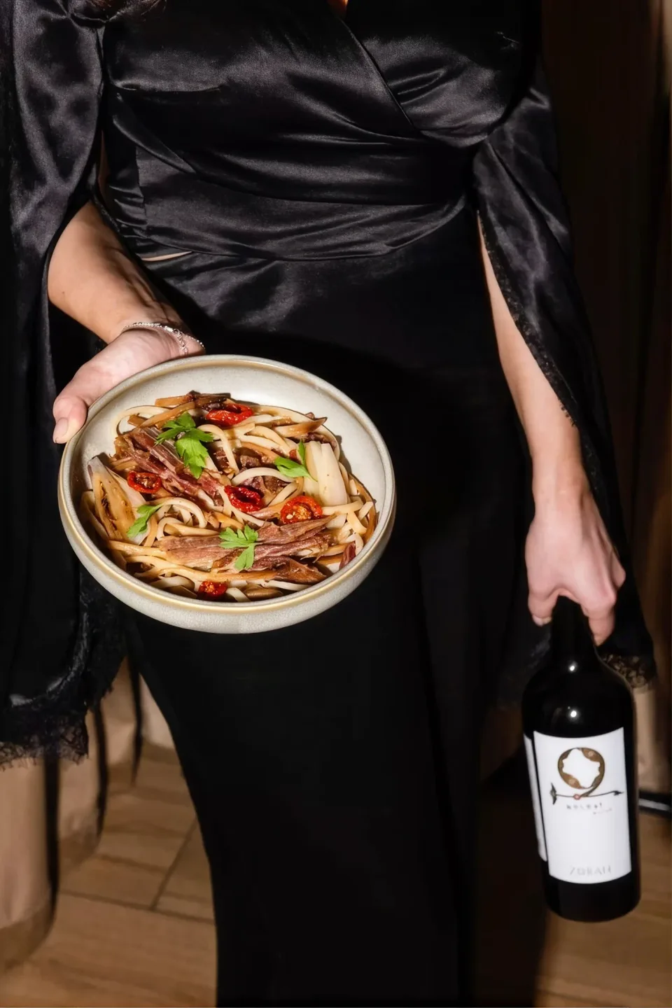 A server in black carrying a bowl of pasta and a bottle of wine