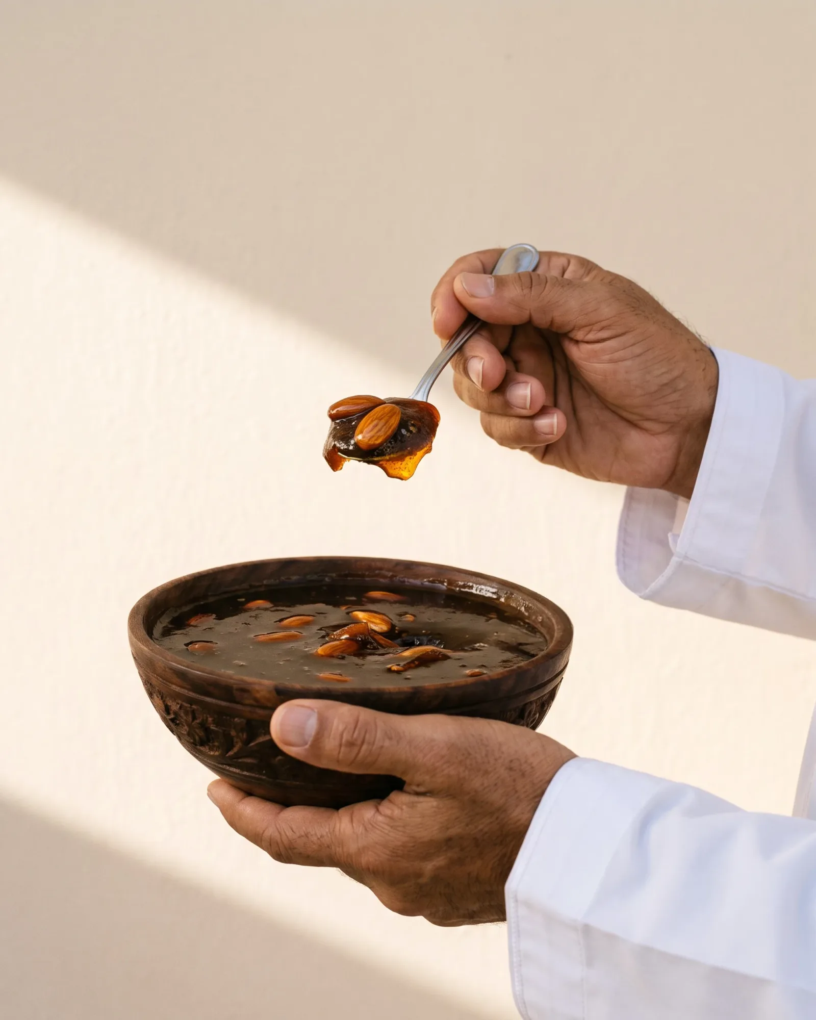 A pair of hands holding a carved wooden bowl of Omani halwa, a spoon lifting a piece clear of the surface, against a warm cream wall.