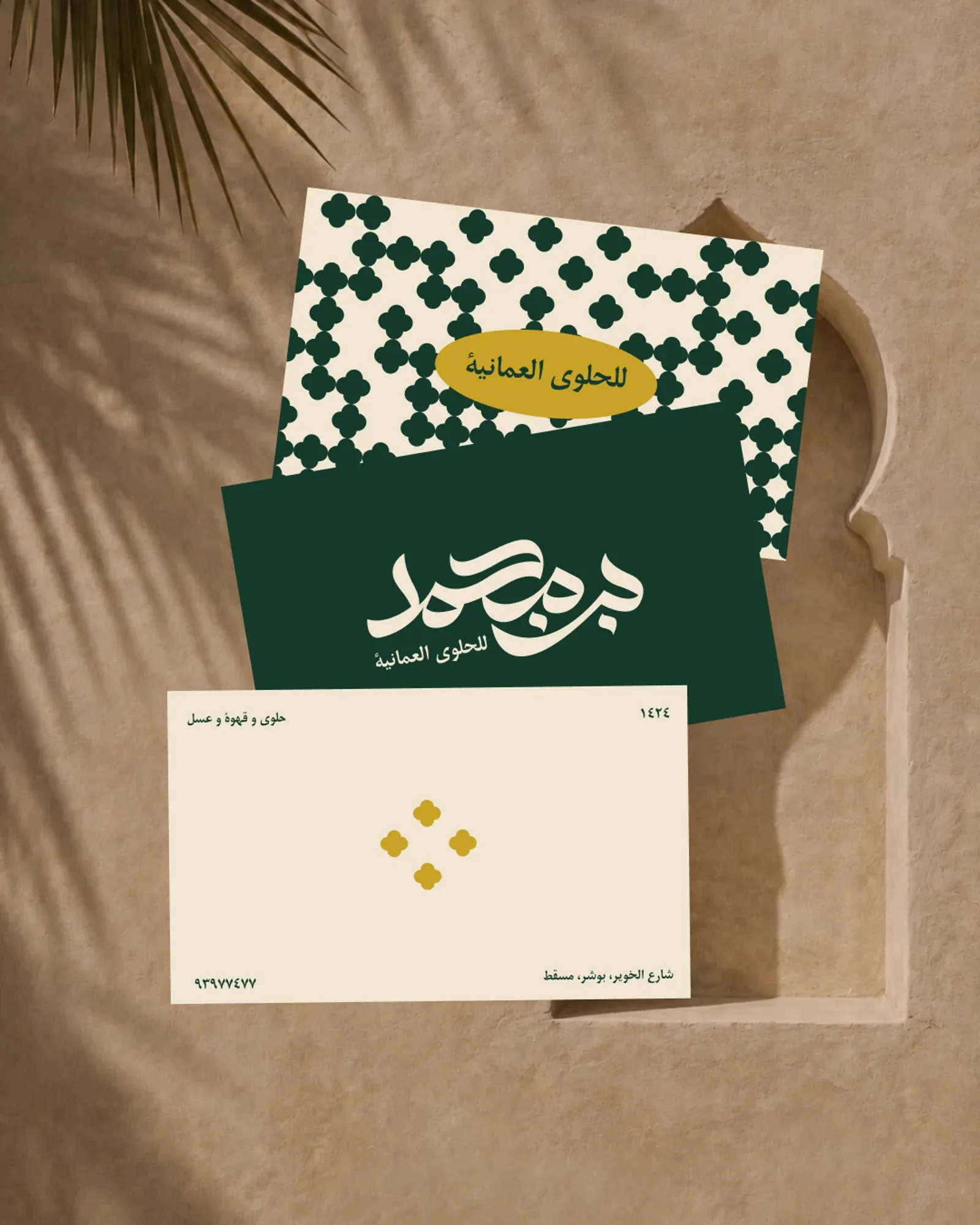 The three business card faces on adobe: the pattern face, the deep green logo face and the cream contact face, palm shadow across them.