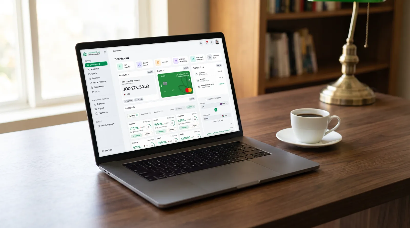 Photograph of a MacBook on a walnut desk beside a coffee cup, the corporate dashboard on its screen: account balance, cards, transactions and the pending approvals.