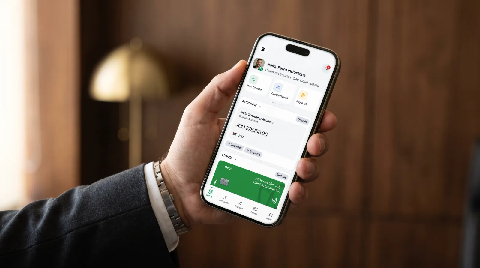 Photograph of a hand holding an iPhone 15 Pro in a walnut and brass office, the corporate banking home screen on the display: the balance of the main operating account and the bottom navigation, every pixel the real product.