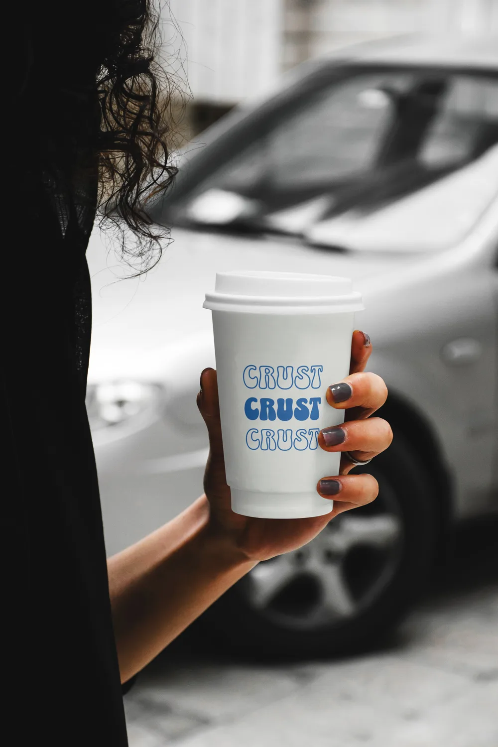 A hand holding a white CRUST coffee cup with the blue mark, photographed against a dark background