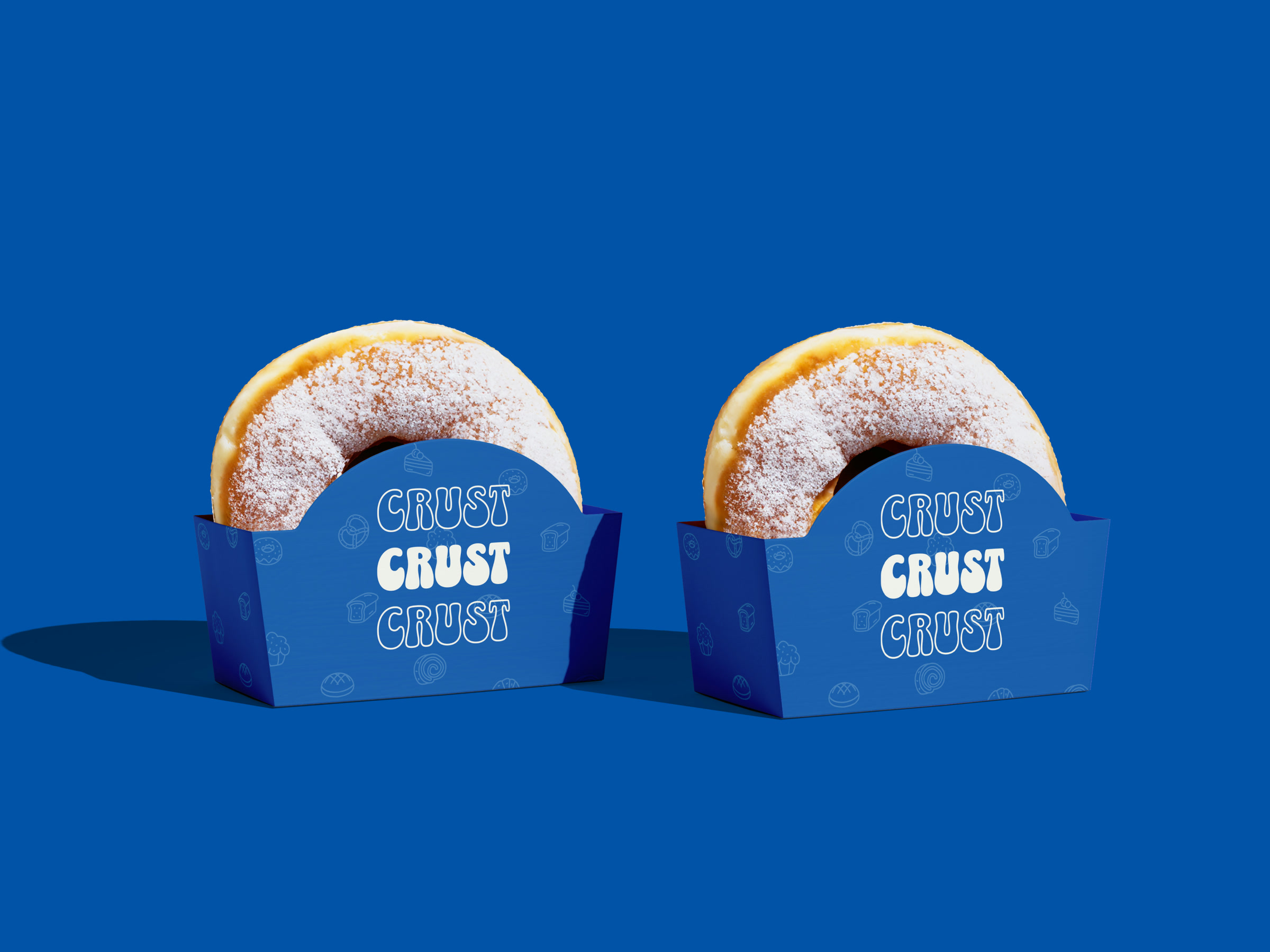 Two blue CRUST doughnut trays on a blue ground, each holding a sugared doughnut above the arched face