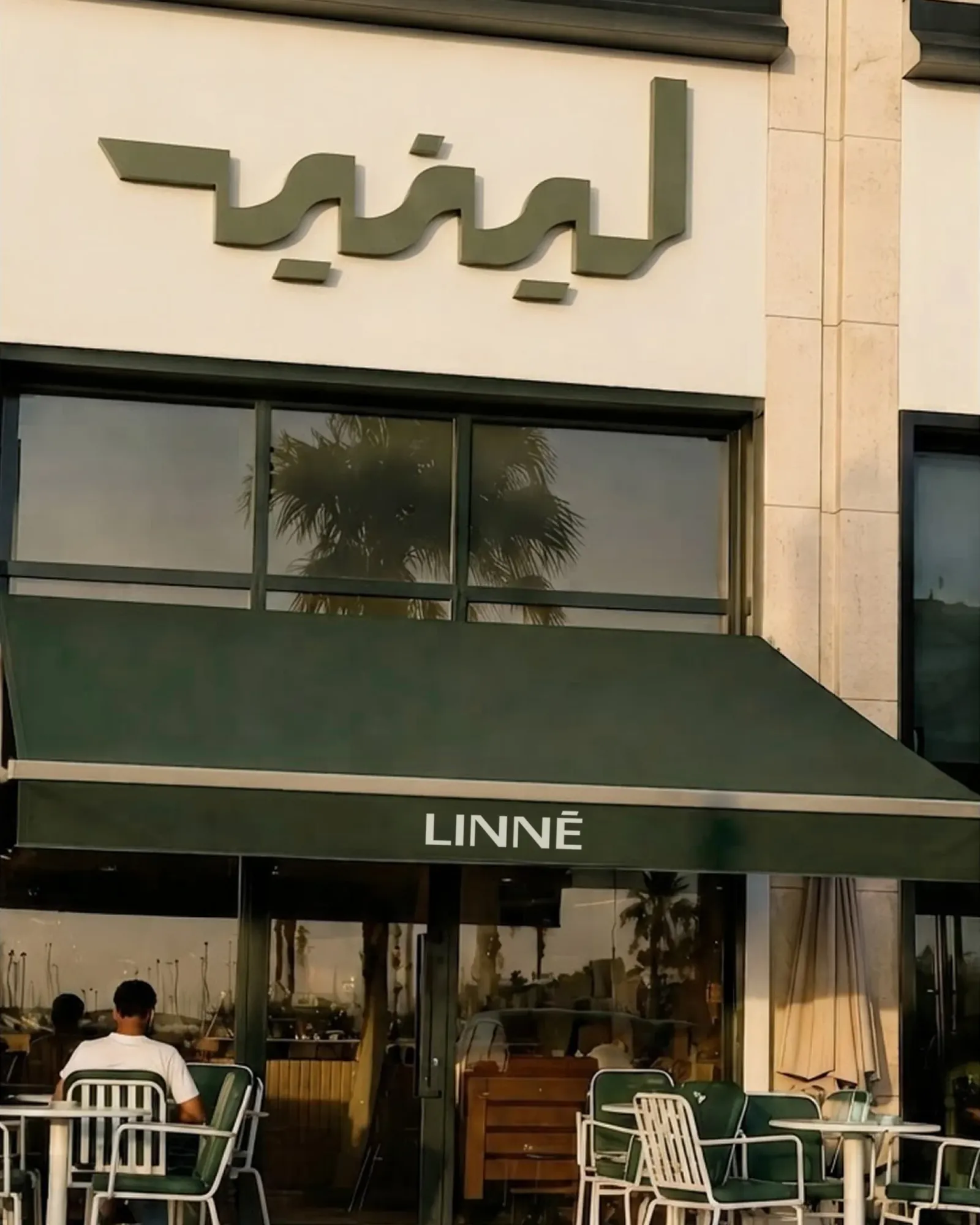 The LINNÉ café front: the Arabic wordmark in relief on the stone above the door, the Latin wordmark on a dark olive awning, a terrace of chairs below.