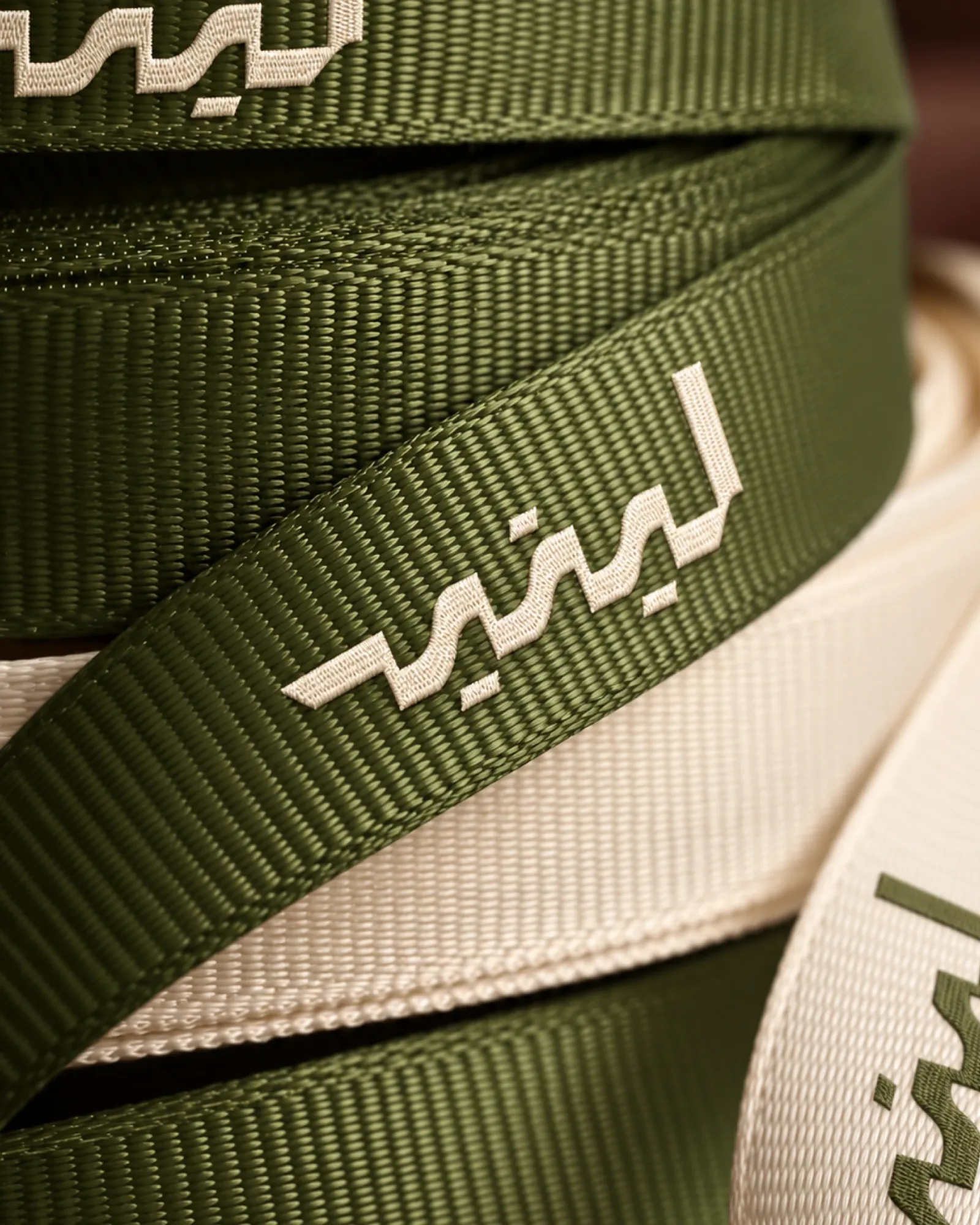 Olive and cream ribbons looped close up, the Arabic wordmark woven into them.