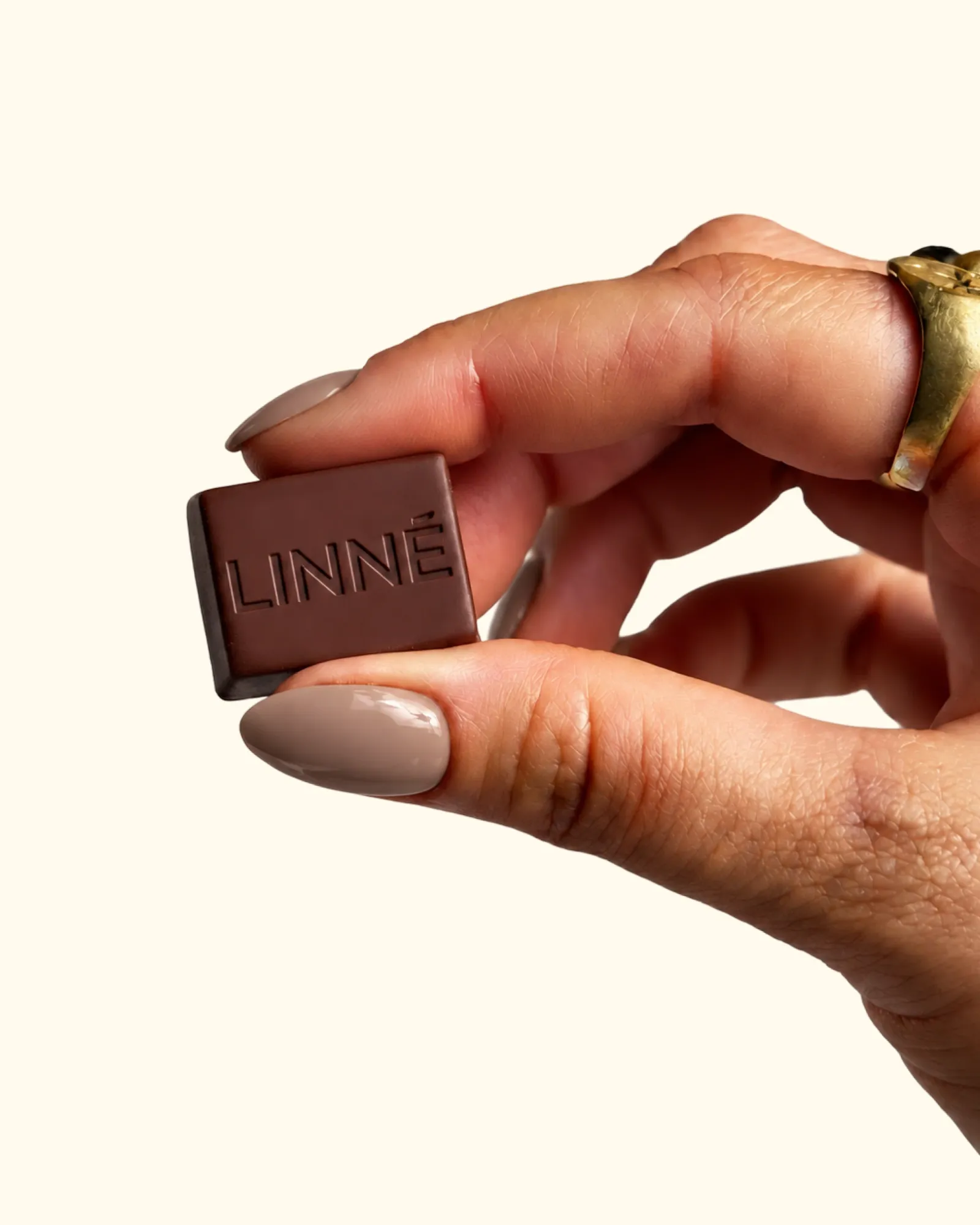 A hand with a gold ring holding a square chocolate pressed with the LINNÉ wordmark, against cream.