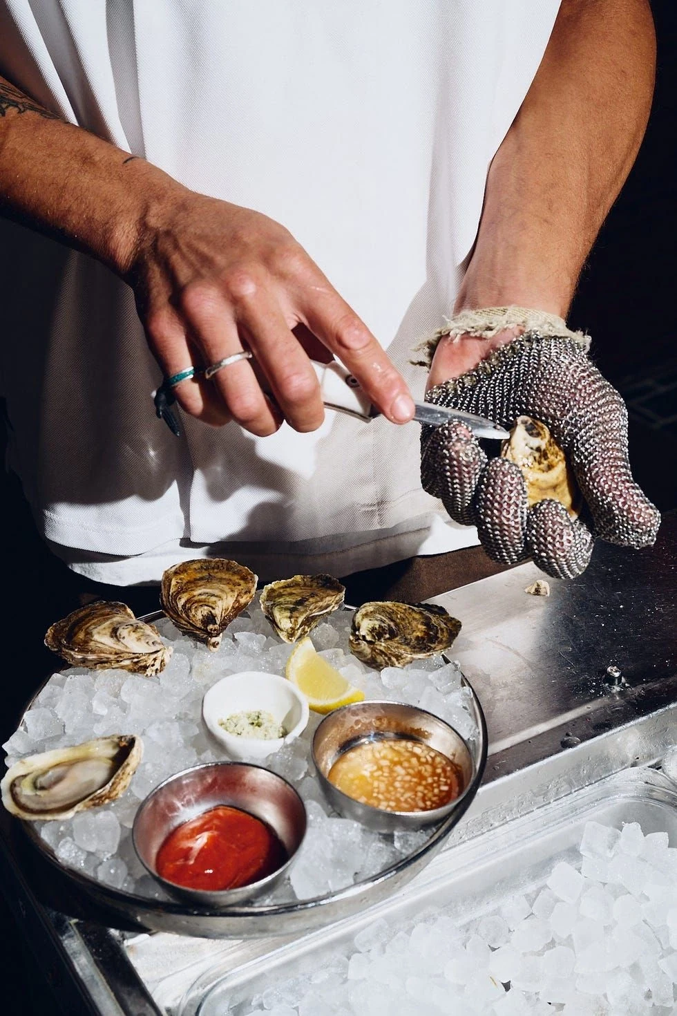 A shucker in a chain-mail glove opening an oyster over a tray of ice, three sauce pots and a lemon wedge beside it