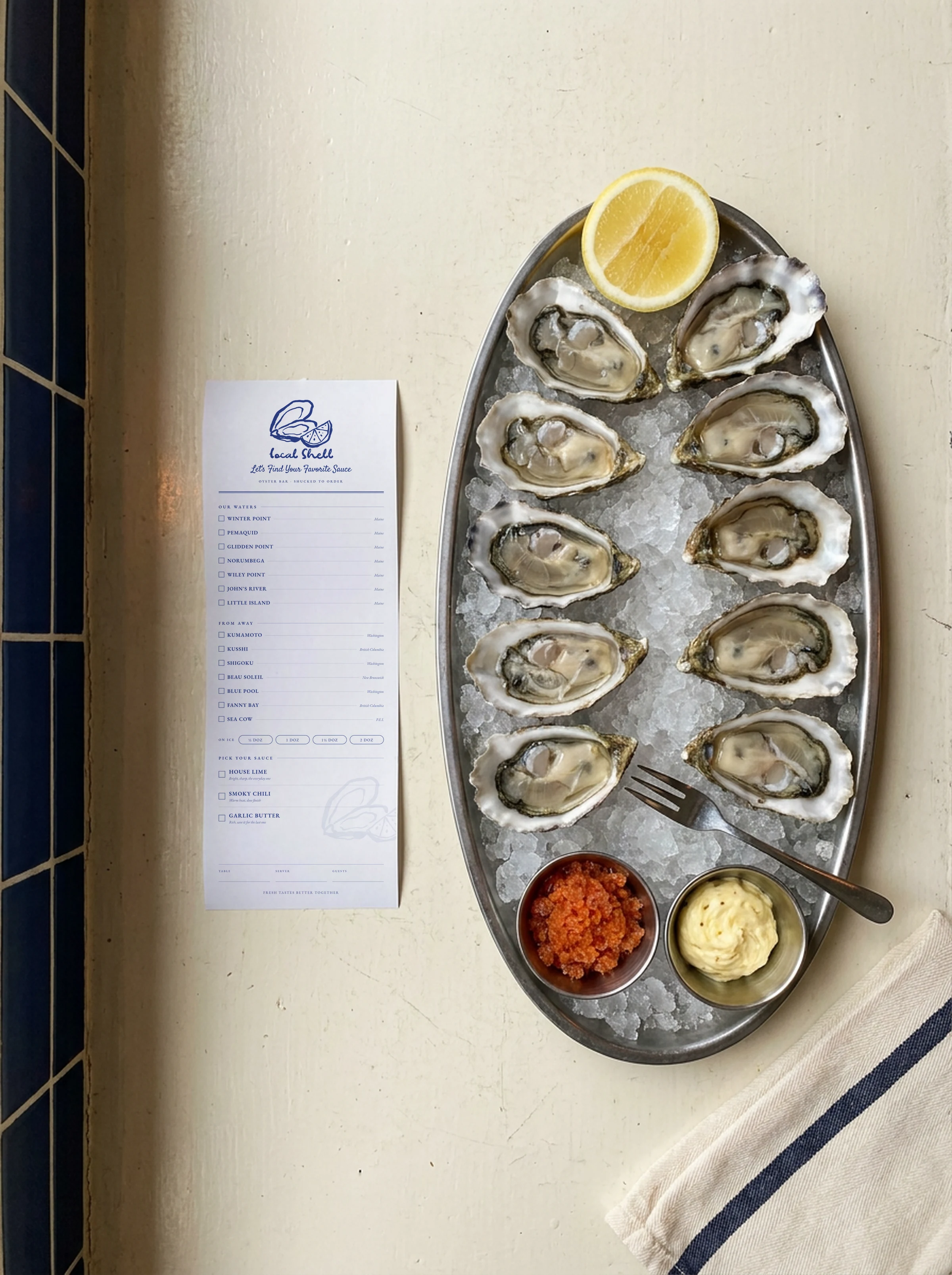 An oval steel platter of a dozen oysters on ice with a lemon half and two sauce pots, on a pale counter beside the printed menu card
