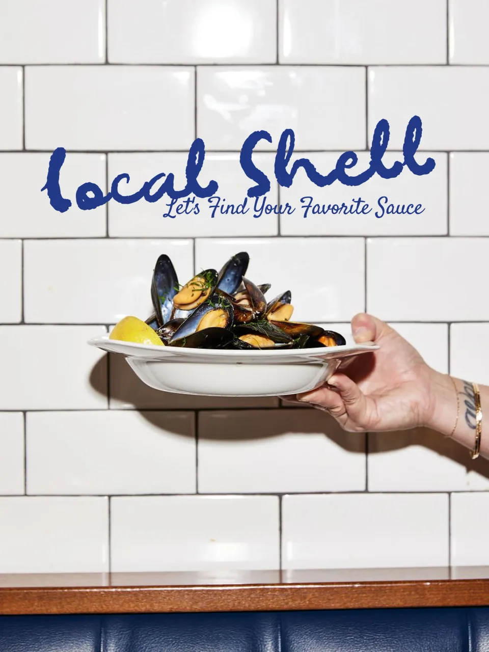 The Local Shell wordmark and its line painted across a white subway-tile wall in brand blue, a hand holding out a bowl of mussels beneath it