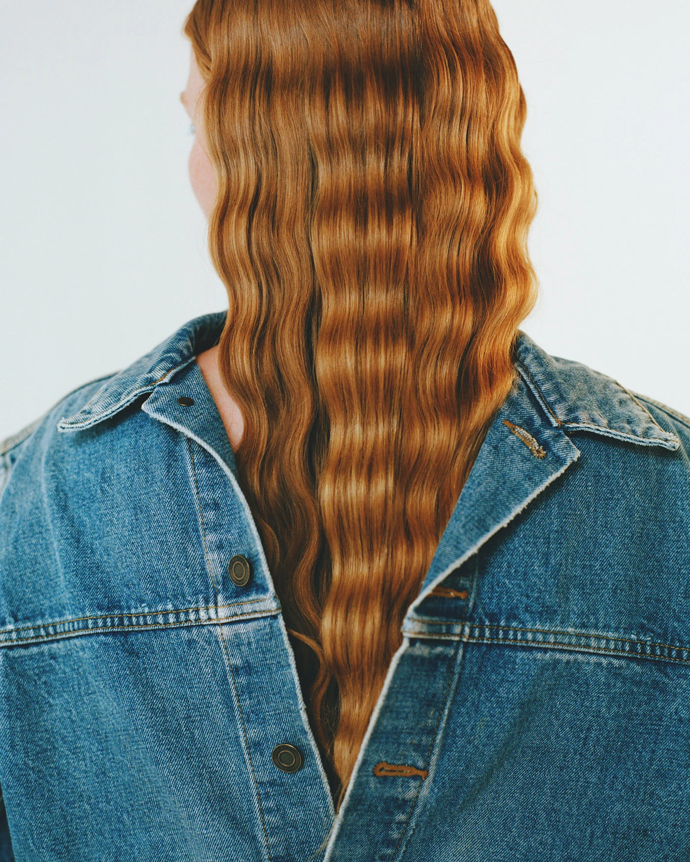 A model seen from behind in a denim jacket, long crimped copper hair falling down the back