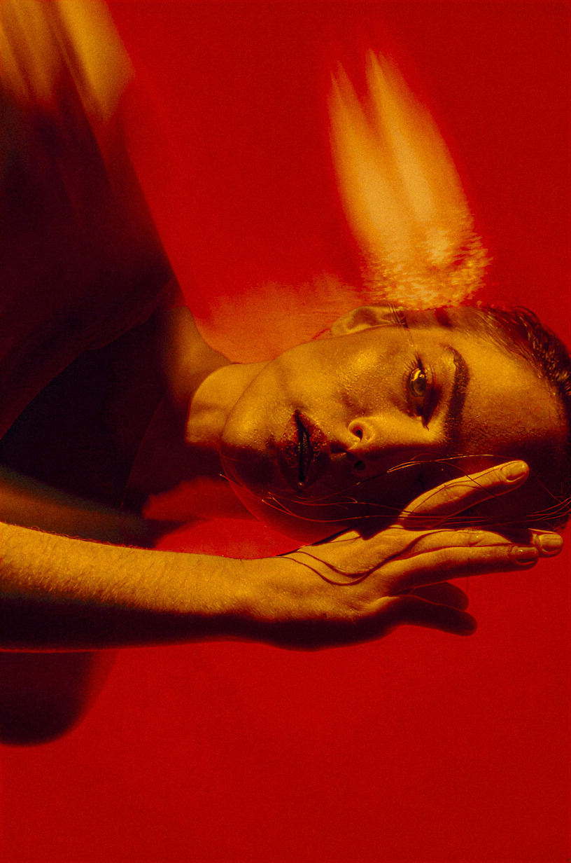 Red gel campaign frame: a model reclining with motion-blurred hair, face resting in an open hand