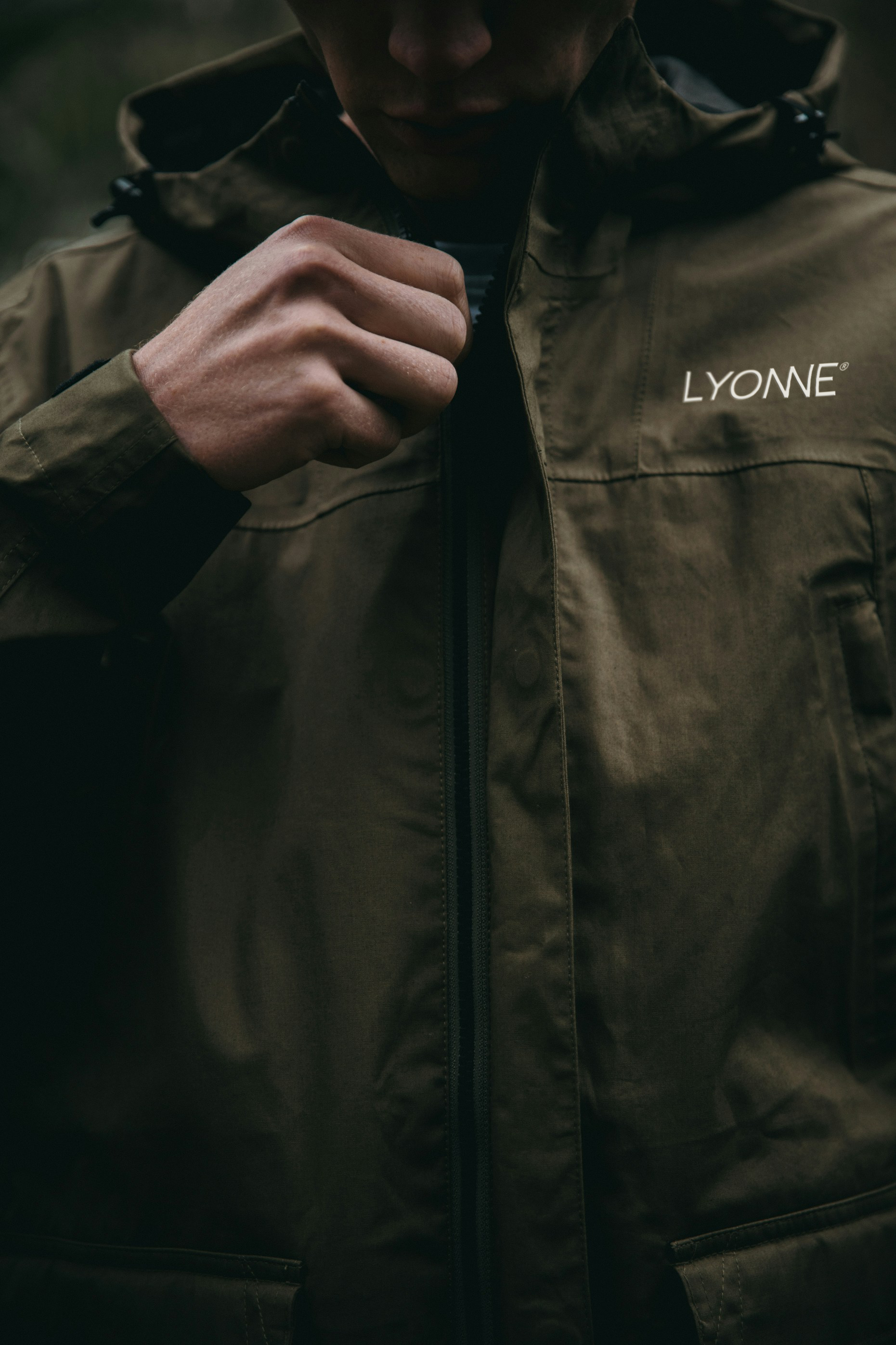 A hand drawing up the zip of an olive technical jacket with the Lyonne wordmark at the chest
