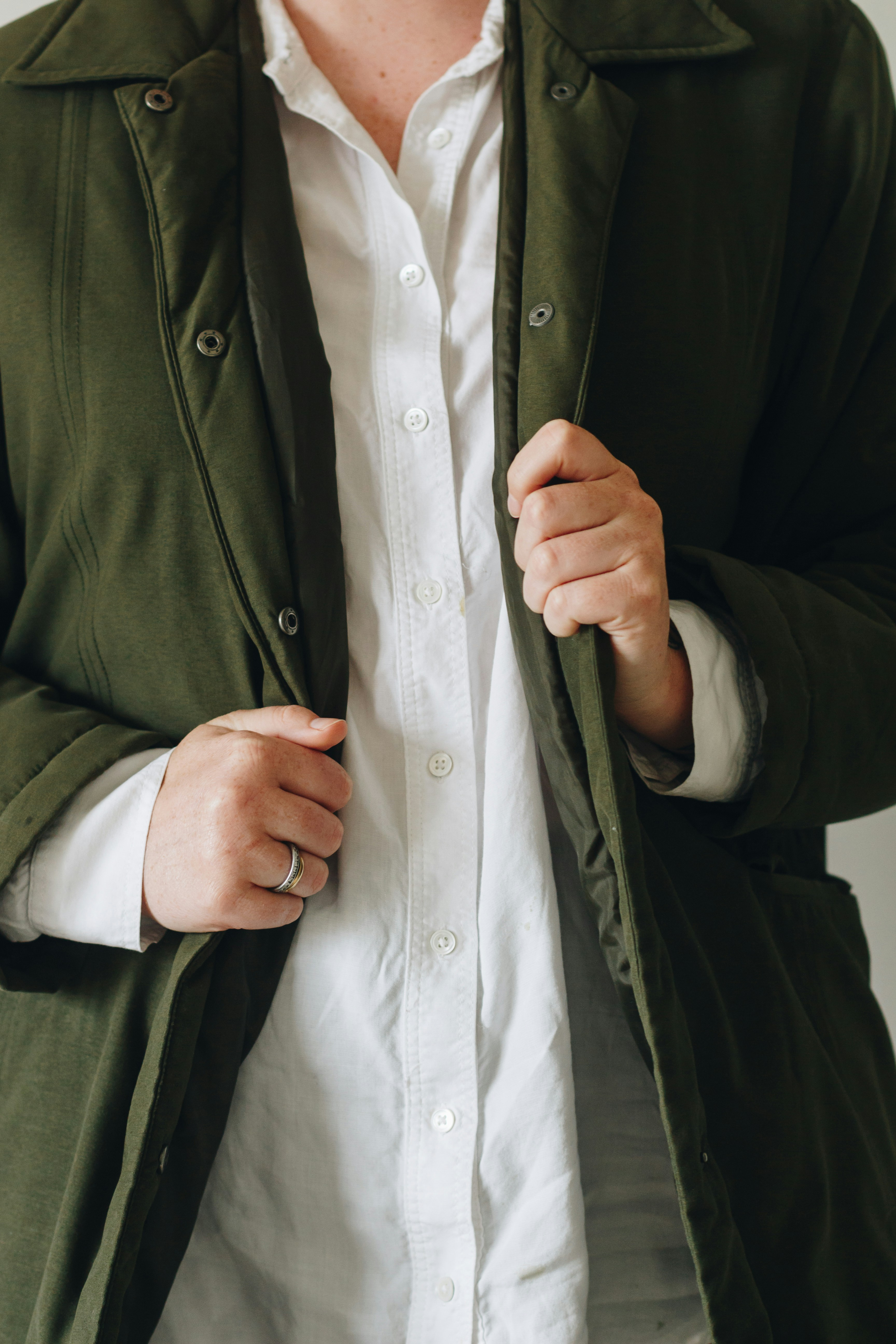 Olive overcoat worn open over a white shirt, hands holding the front edges