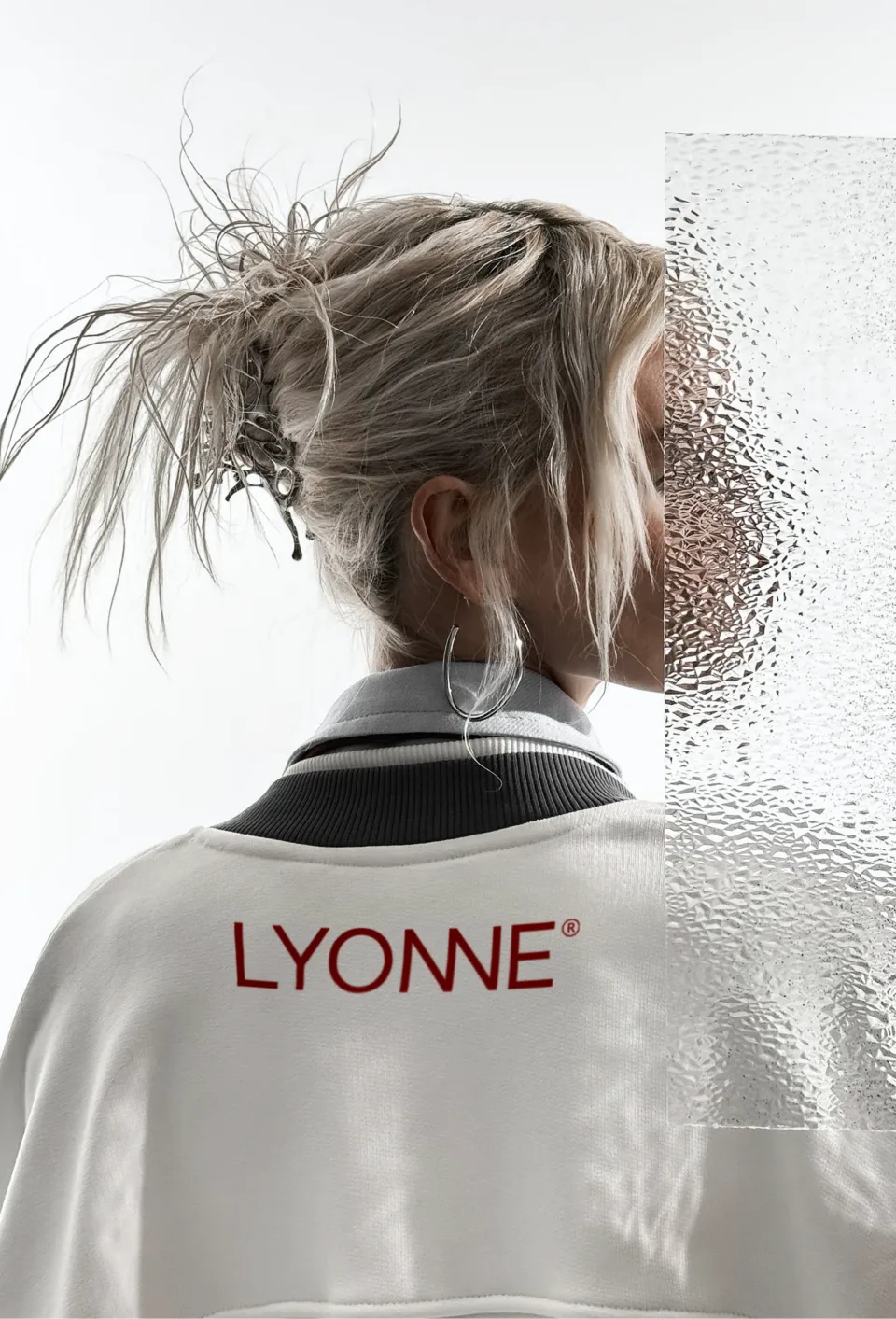 The back of a white Lyonne sweatshirt with the red wordmark, worn by a model with tousled blonde hair, beside a panel of textured glass
