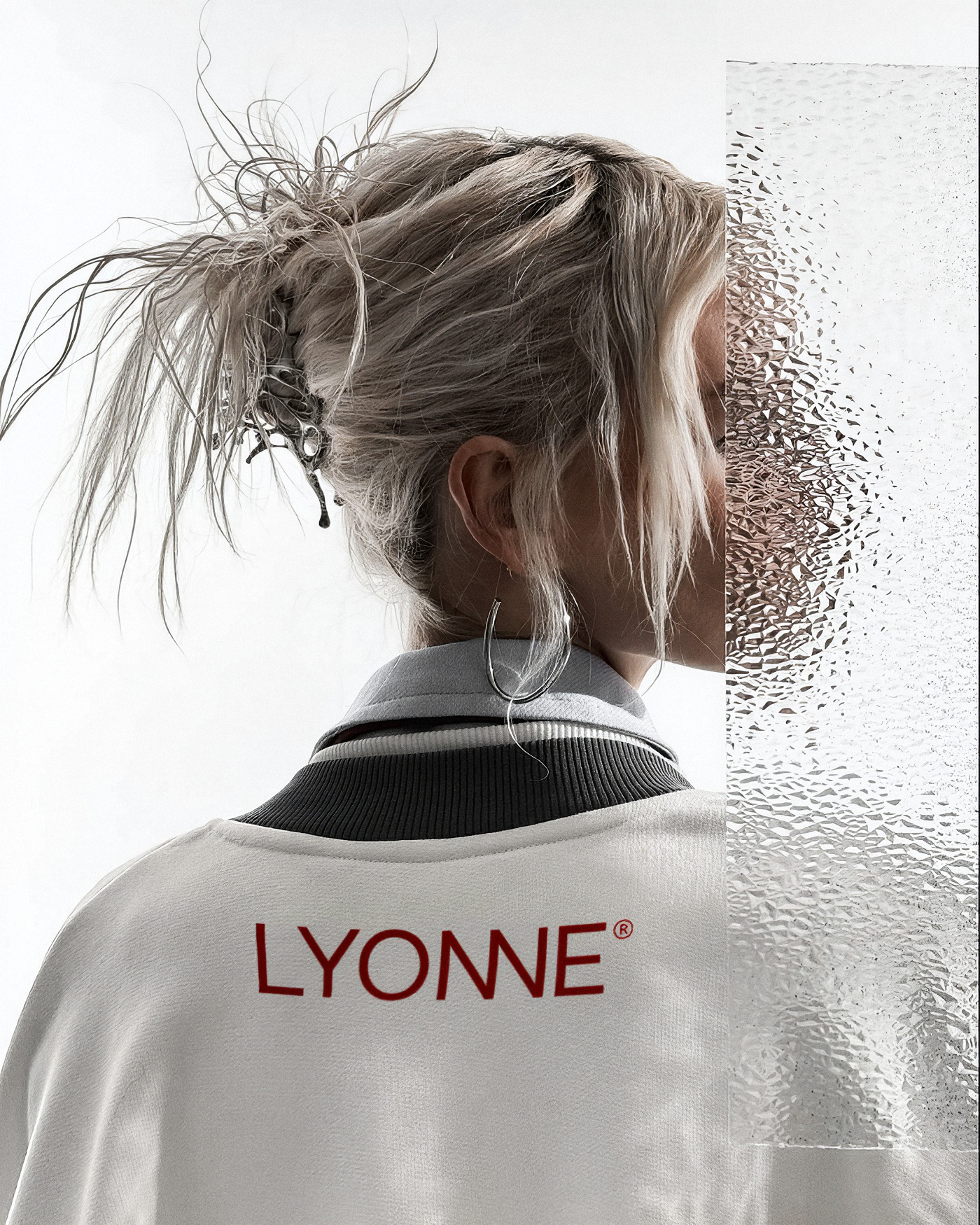 Back of a white Lyonne varsity jacket with the red wordmark, worn by a model with a sculptural updo beside fluted glass