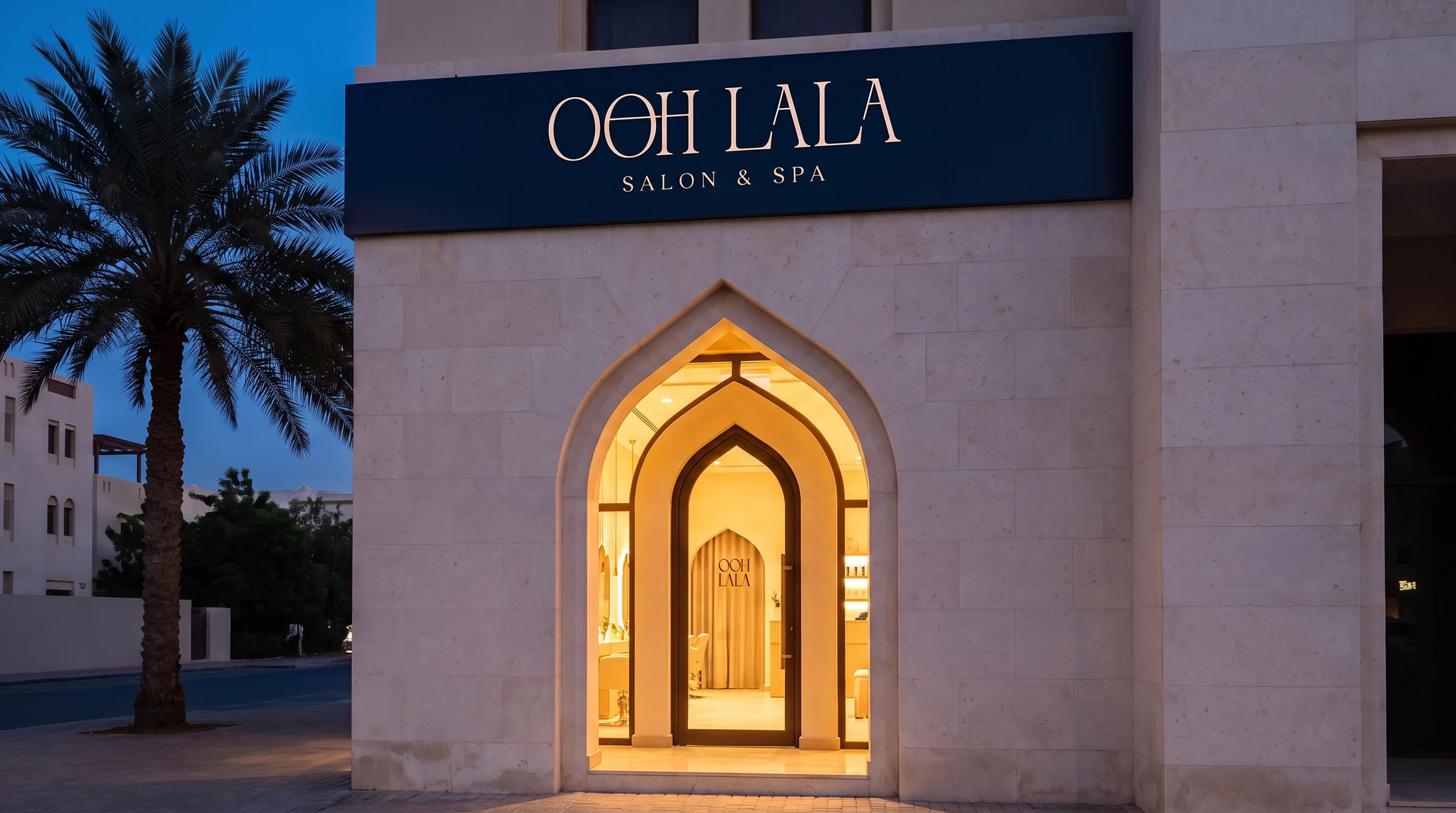 The salon at dusk seen wide: pale stone facade, navy fascia lettered OOH LALA SALON & SPA, a lit pointed arch doorway, palm trees against a deep blue sky