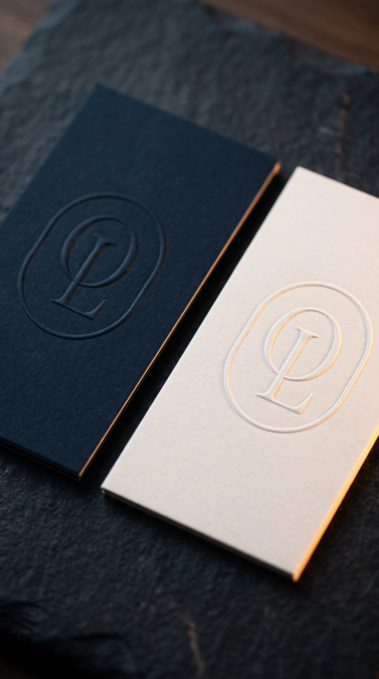 A navy folder and a white card on dark stone, both carrying the monogram, the card blind embossed and the folder foiled