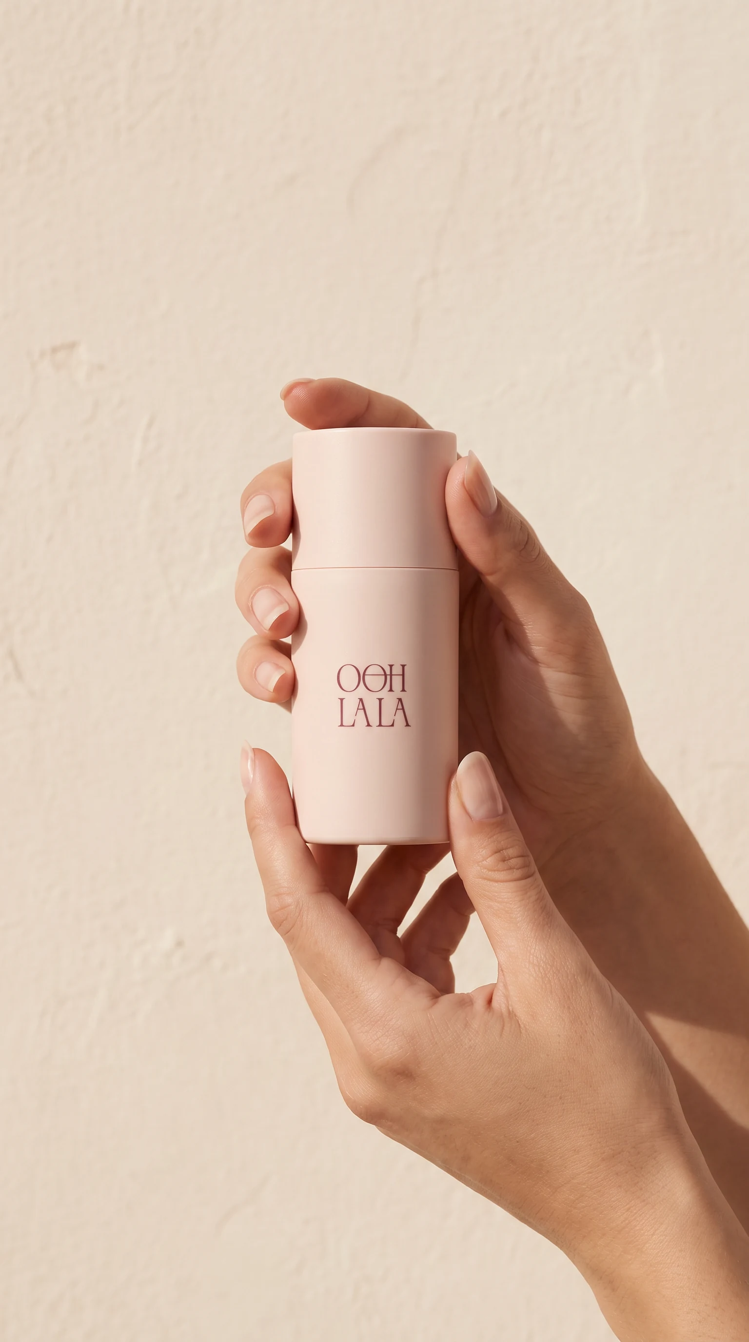 A hand holding a blush cylindrical bottle against a plaster wall, the monogram small at its foot
