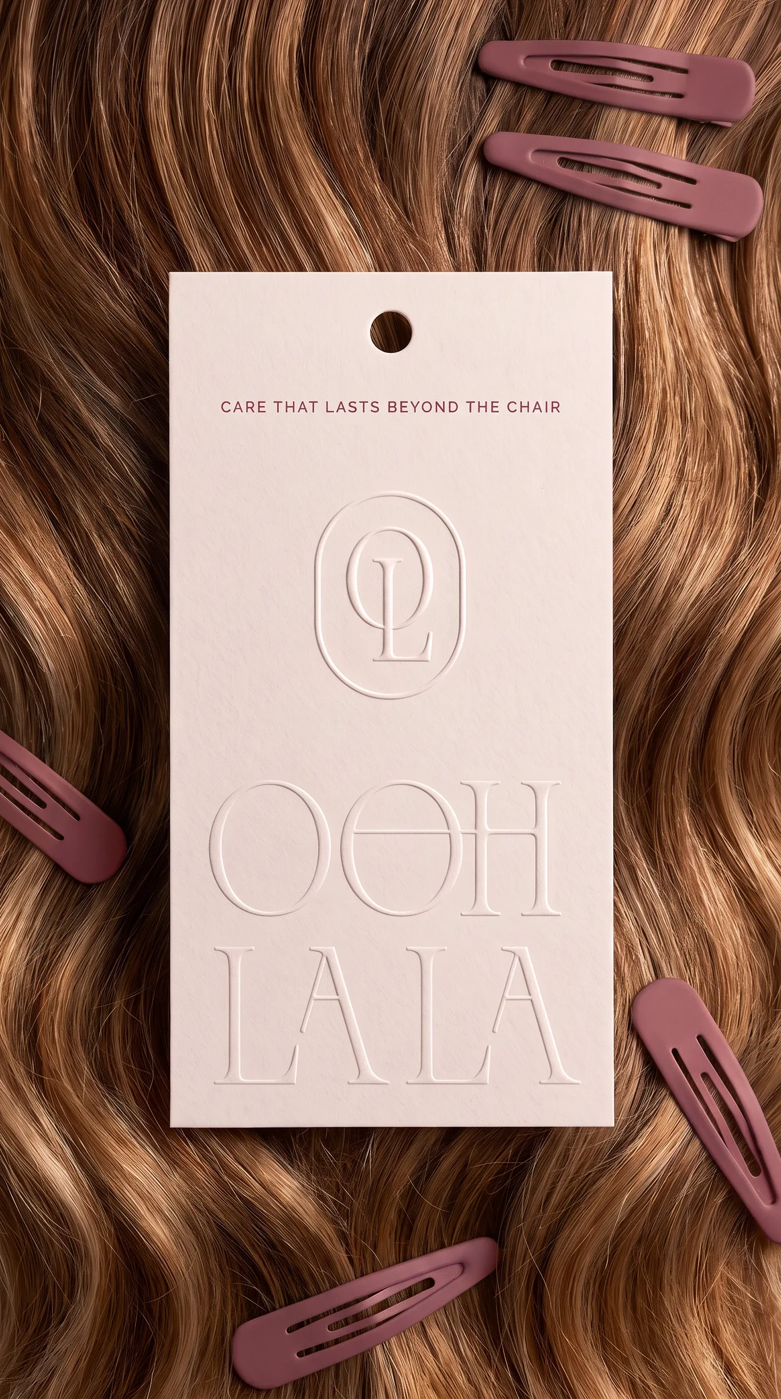 A blush hang tag on auburn waves, embossed with the monogram, the wordmark and the line CARE THAT LASTS BEYOND THE CHAIR, rose hair clips scattered around