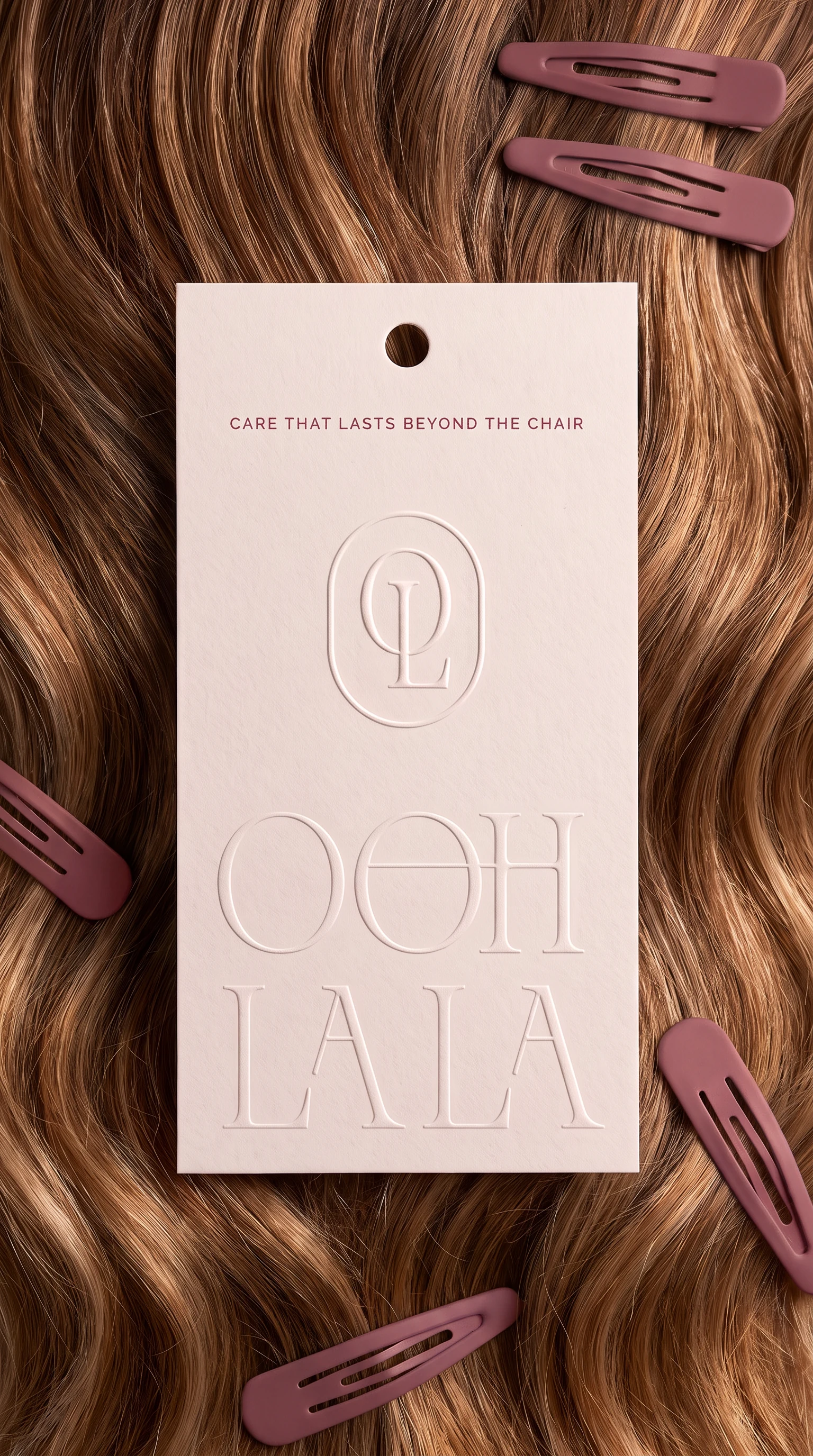 A blush hang tag on auburn waves, embossed with the monogram, the wordmark and the line CARE THAT LASTS BEYOND THE CHAIR, rose hair clips scattered around