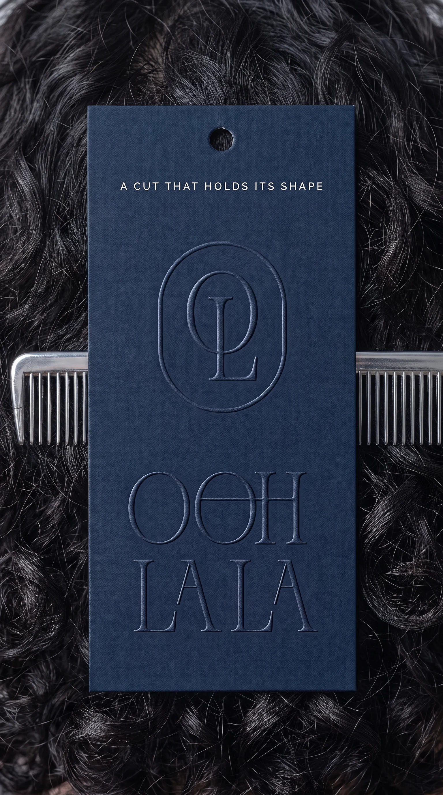 A navy hang tag on dark curly hair, blind embossed with the monogram, the wordmark and the line A CUT THAT HOLDS ITS SHAPE, a comb behind it