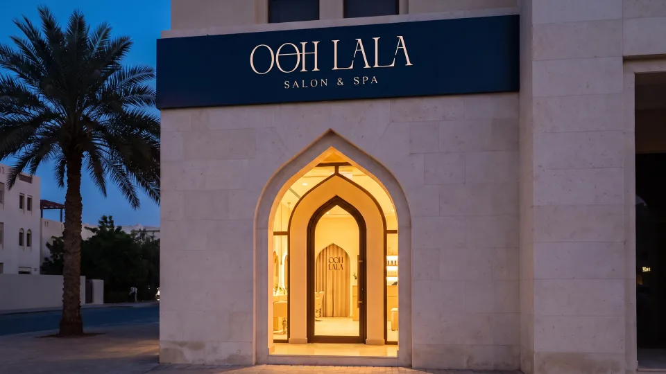 The Ooh Lala shopfront at dusk: a navy fascia carrying the wordmark above a tall pointed arch cut into pale stone, the doorway lit warm gold from inside, palms either side