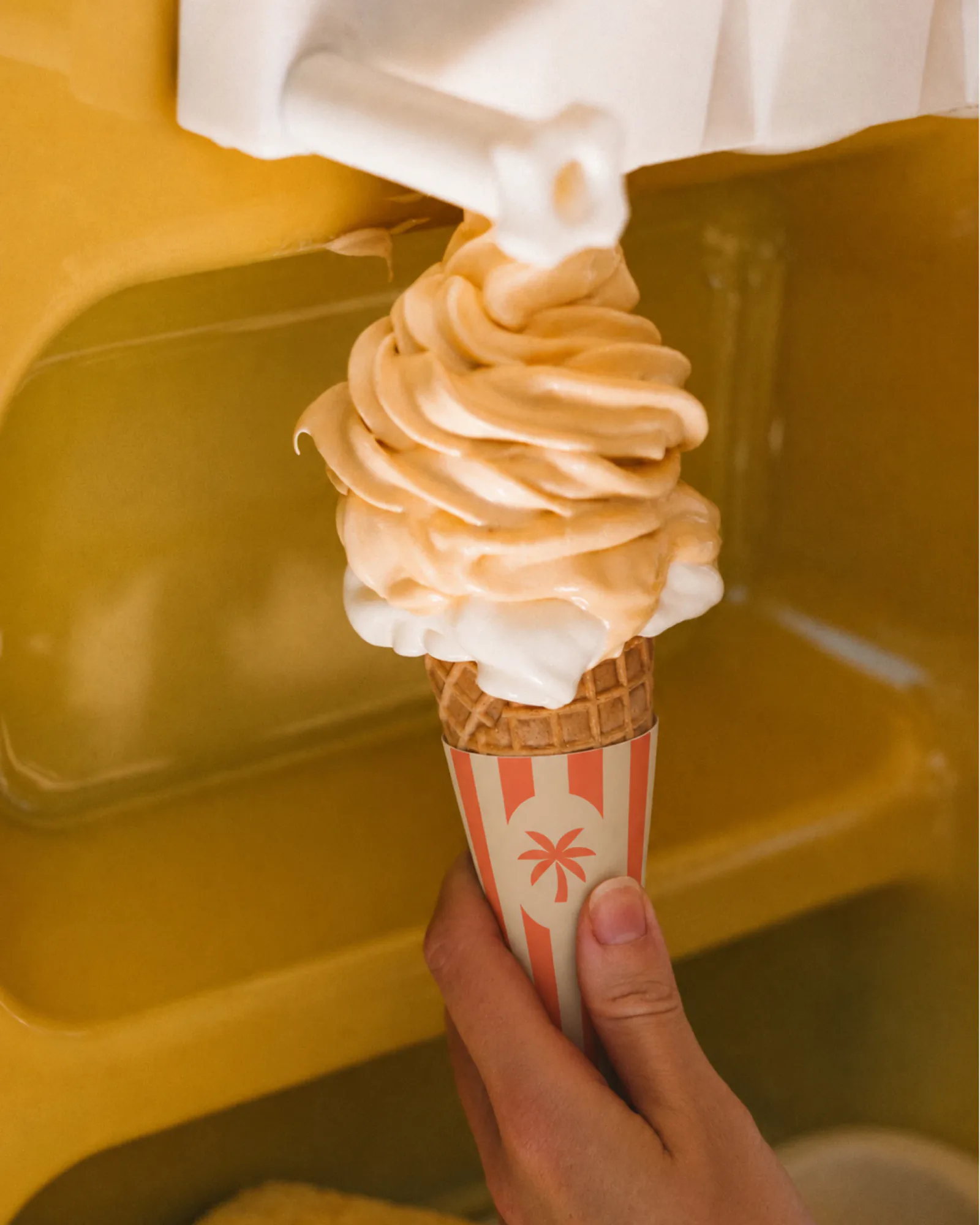Soft serve pouring onto a cone in a striped palm sleeve, against a yellow machine.