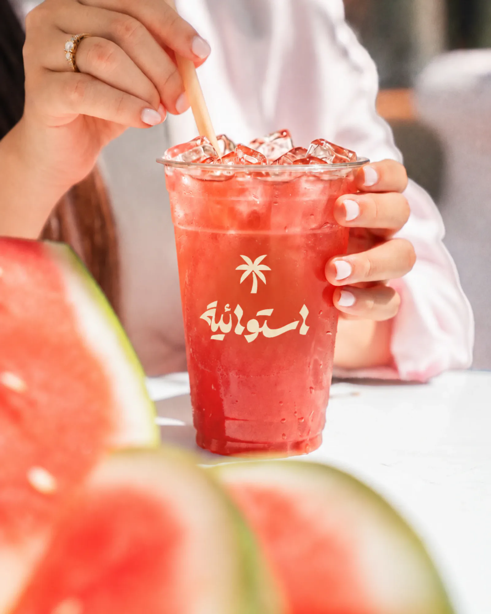 An iced watermelon juice in a cup printed with the palm and the Arabic wordmark, watermelon slices in front.