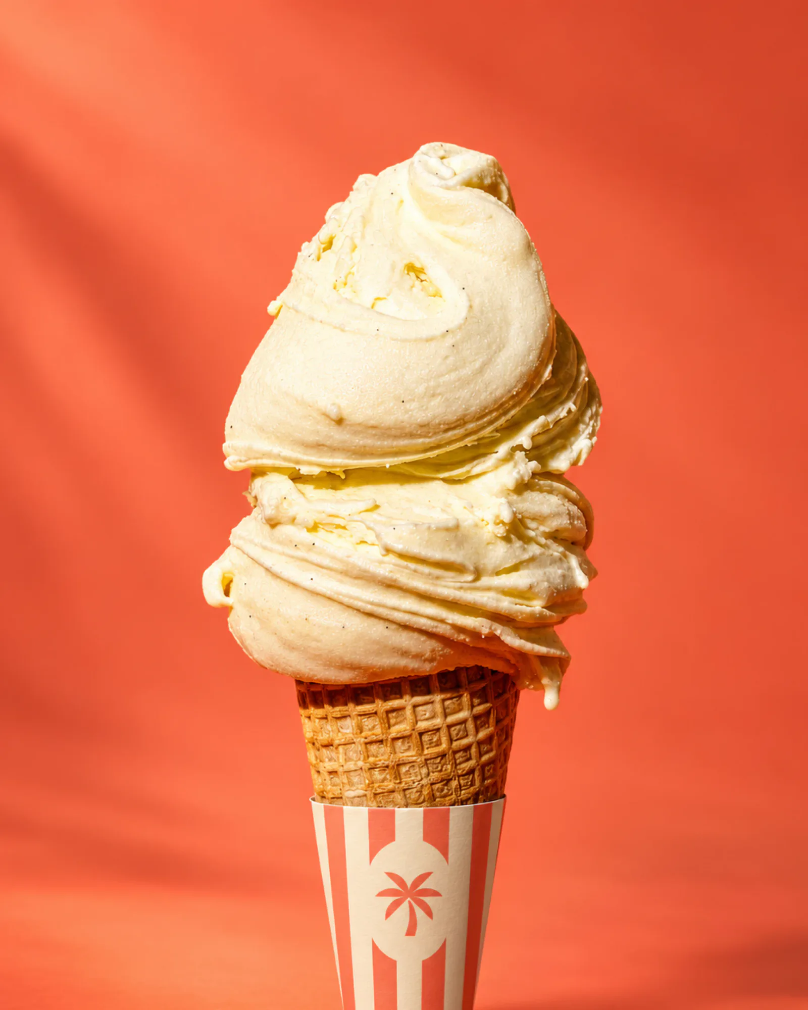 A scoop of vanilla ice cream on a waffle cone in a coral and cream striped sleeve with the palm, against a coral wall.