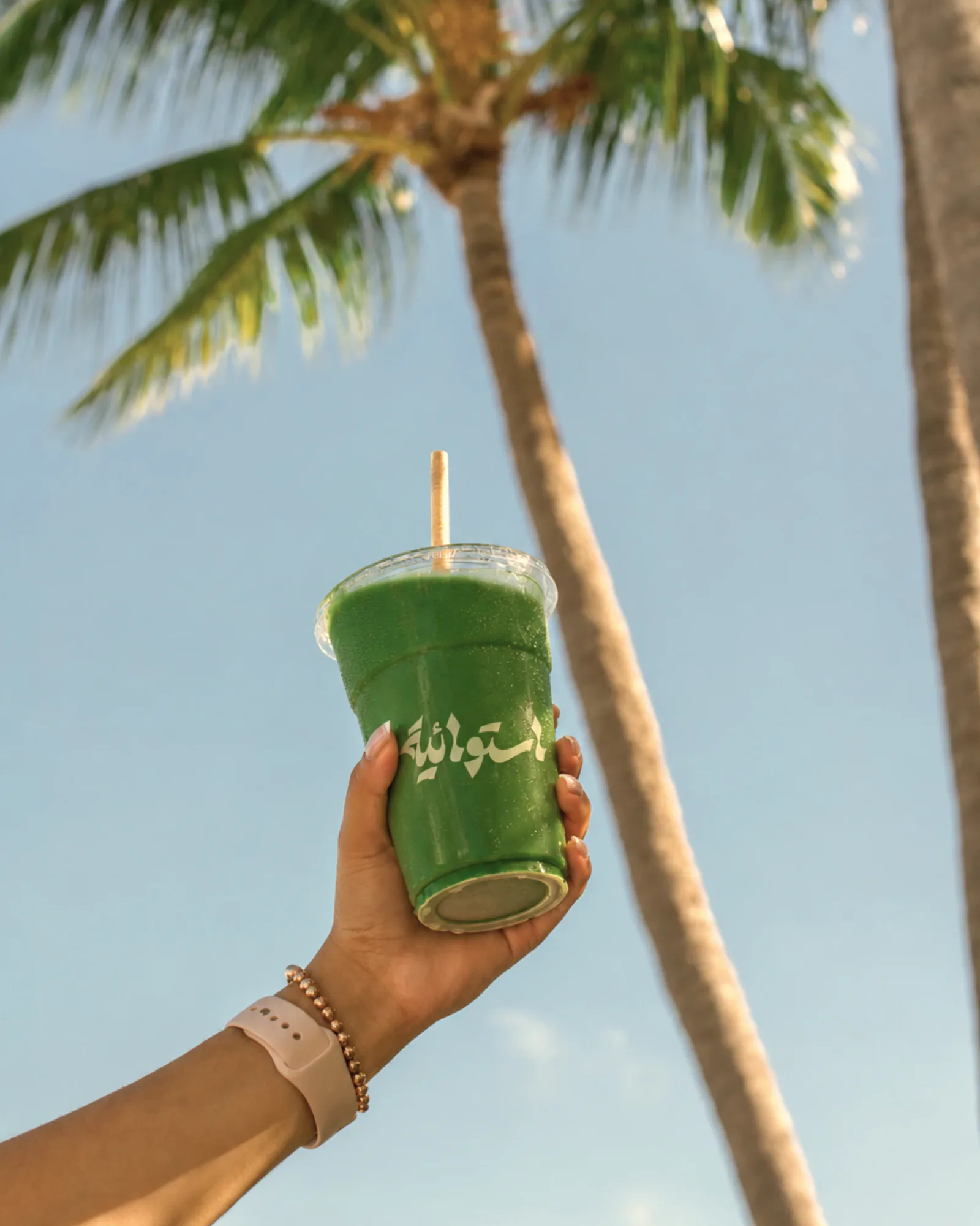 A hand holding up a green juice in a clear cup printed with the Arabic wordmark, under a palm tree and blue sky.
