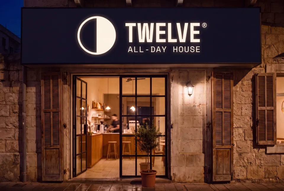 The same TWELVE shopfront at night, the sign panel now inverted to near-black with the mark and wordmark glowing bone out of it, over a wet street
