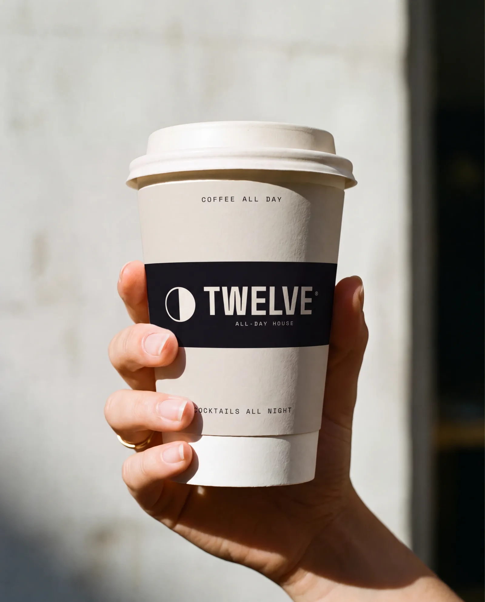 A hand holding a TWELVE takeaway cup in hard morning sun, the dark printed band wrapping around the paper cylinder