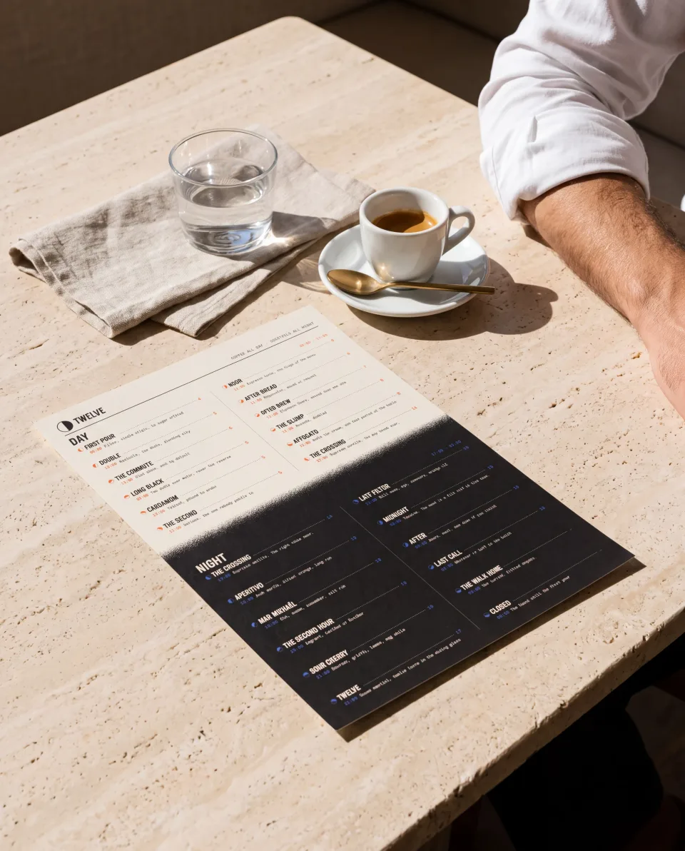The TWELVE all-day menu card on a travertine cafe table in morning sun, its day half on bone above the sprayed crossing and its night half on ink below