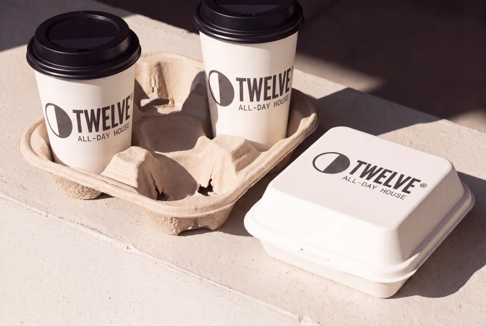 A pulp four-cup carrier holding two bone TWELVE takeaway cups beside a moulded clamshell box, on a sunlit concrete ledge, every piece printed dark on bone